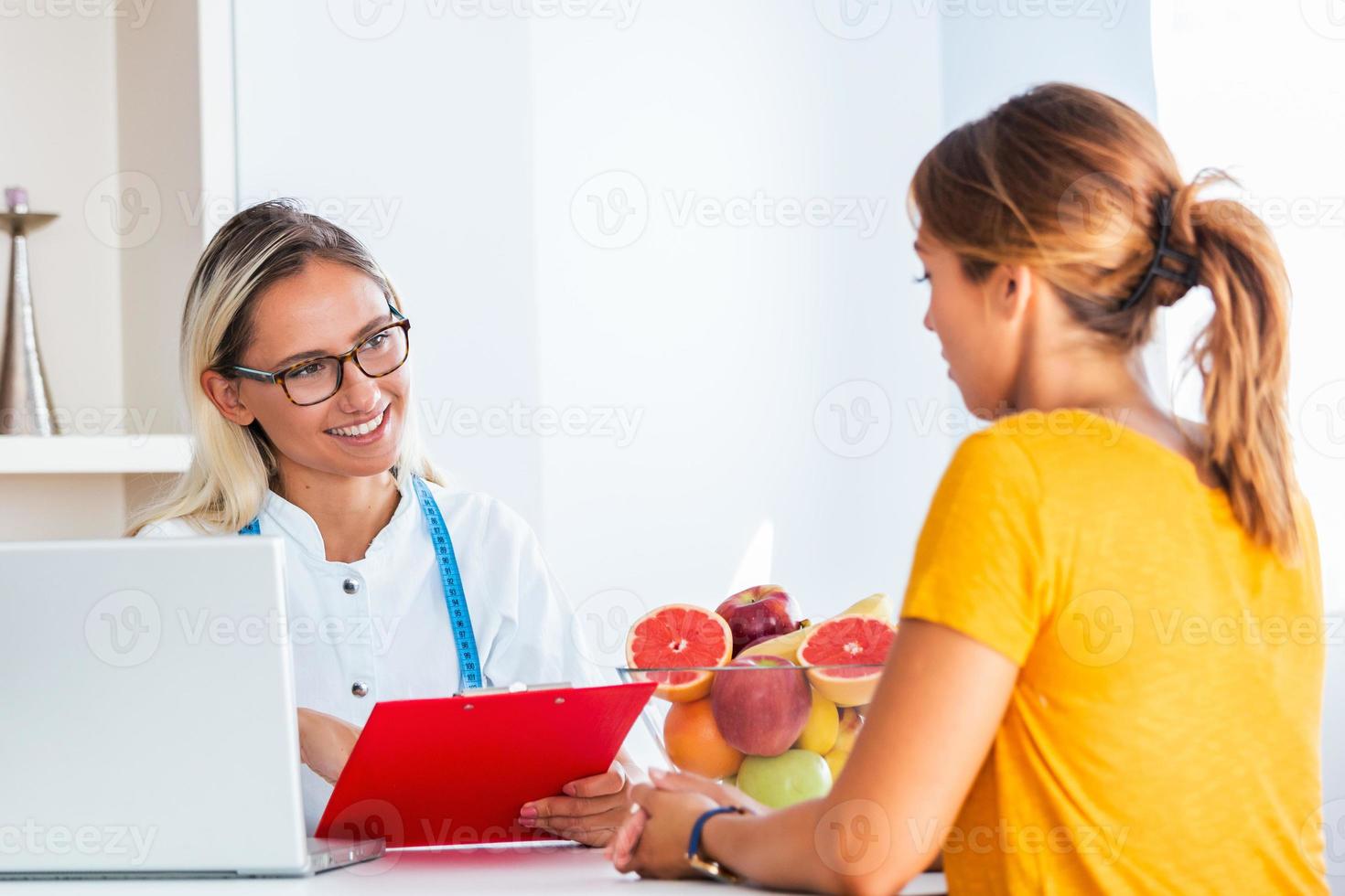 dokter voedingsdeskundige, diëtist en vrouw geduldig Aan overleg in de kantoor. jong glimlachen vrouw voedingsdeskundige in de overleg kamer. voedingsdeskundige bureau met gezond fruit en meten plakband. foto