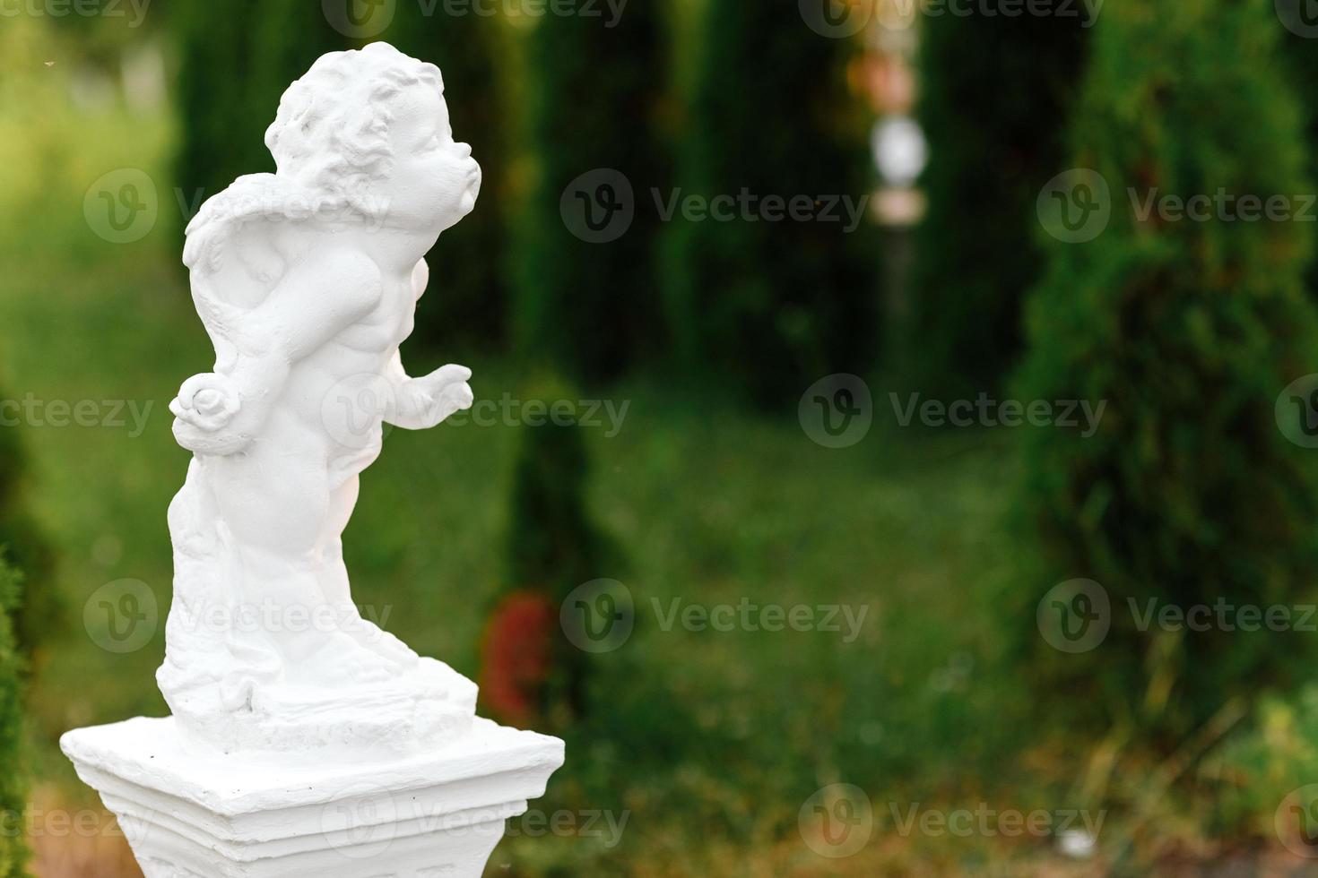 standbeeld van baby engel met Vleugels tegen groen tuin. de schattig weinig Cupido engel beeldhouwwerk standbeeld staand buitenshuis foto