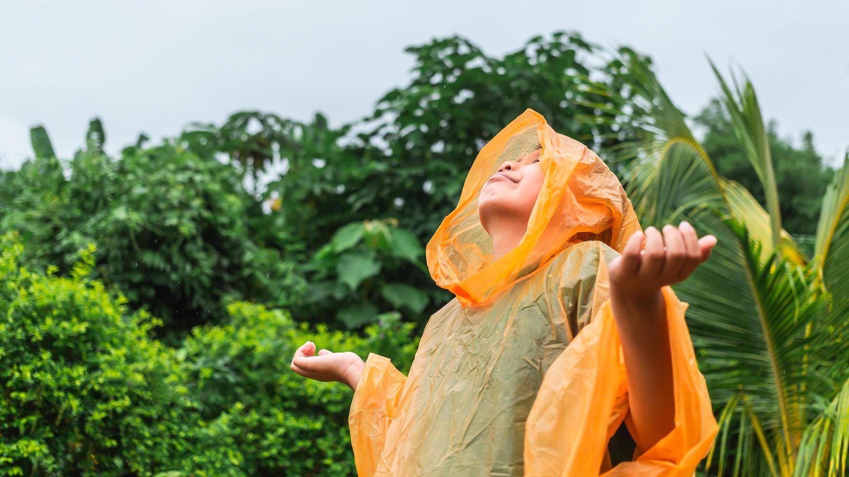 Aziatisch jongen vervelend oranje regenjas is gelukkig en hebben pret in de regen Aan een regenachtig dag. foto