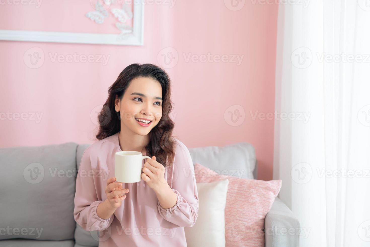 vrolijk jong Aziatisch vrouw Holding een mok wanneer zittend Aan sofa Bij huis. foto