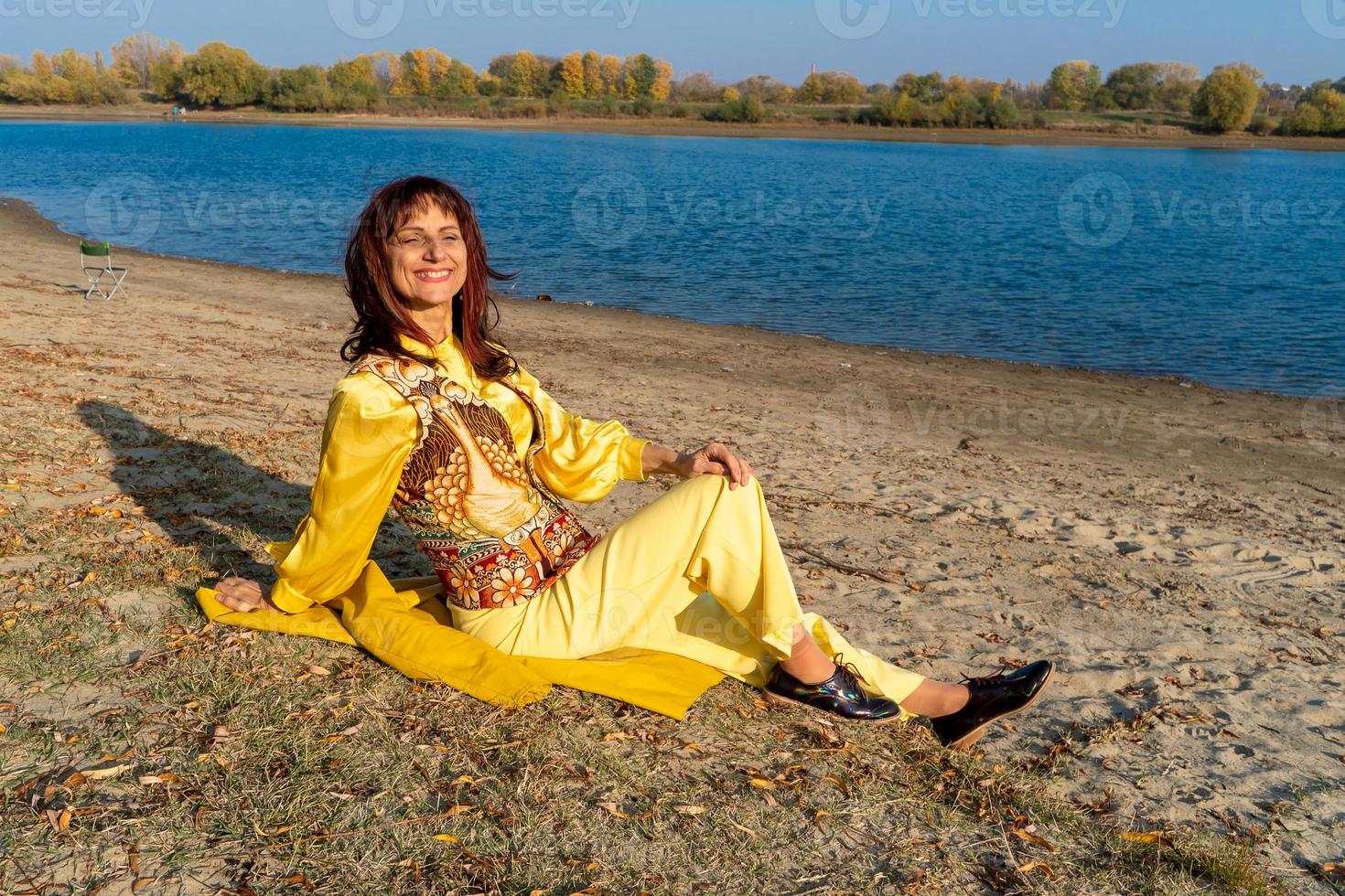 een vrouw in geel kleren en een elegant hesje is zittend door de meer. positief leven concept foto