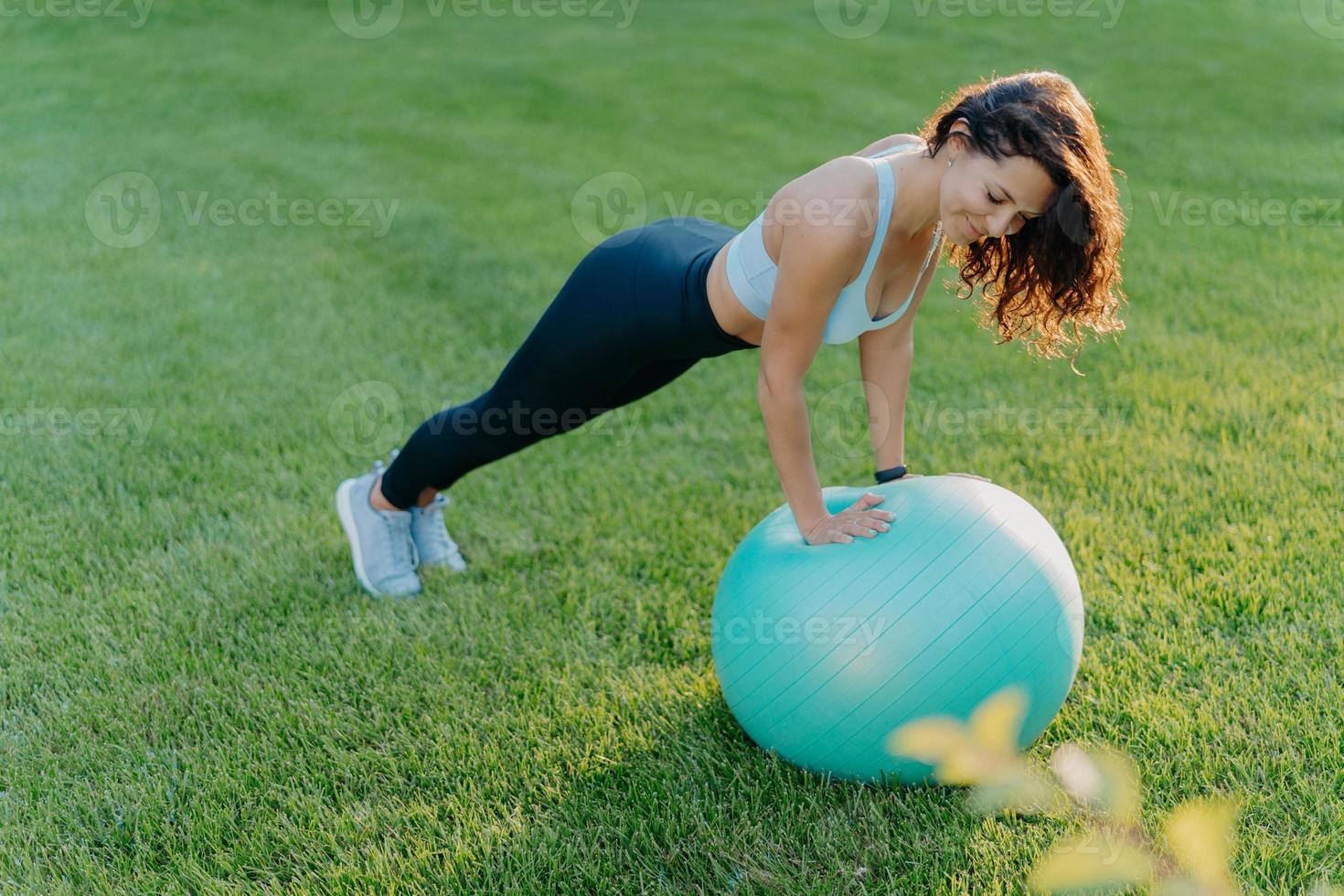 sportief meisje staat in plank pose op gymnastiekbal, gekleed in sportkleding, doet fysieke oefeningen op groen gazon buiten, blijft fit en gezond. mensen, yoga, flexibiliteit, lifestyle concept. foto