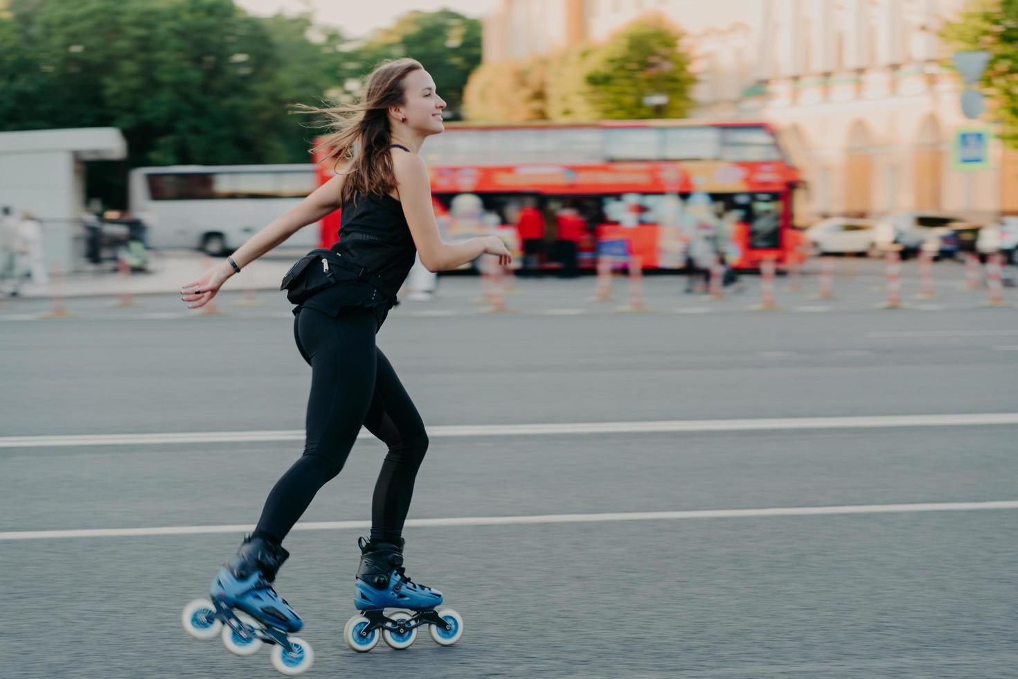 buiten schot van actieve slanke jonge vrouw geniet van rolschaatsen tijdens vrije tijd gekleed in blackactive wear poses in stedelijke plaats op weg tegen onscherpe achtergrond met vervoer. hobby concept foto