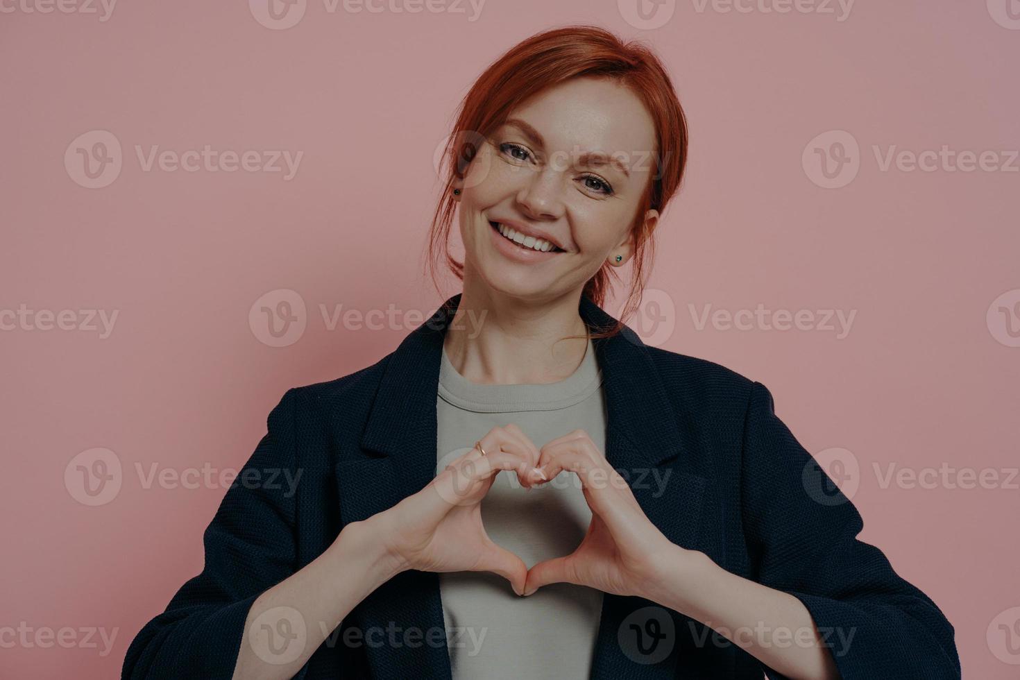 geïsoleerde studio-opname van een jonge, glimlachende roodharige vrouw die met beide handen hartvorm maakt foto