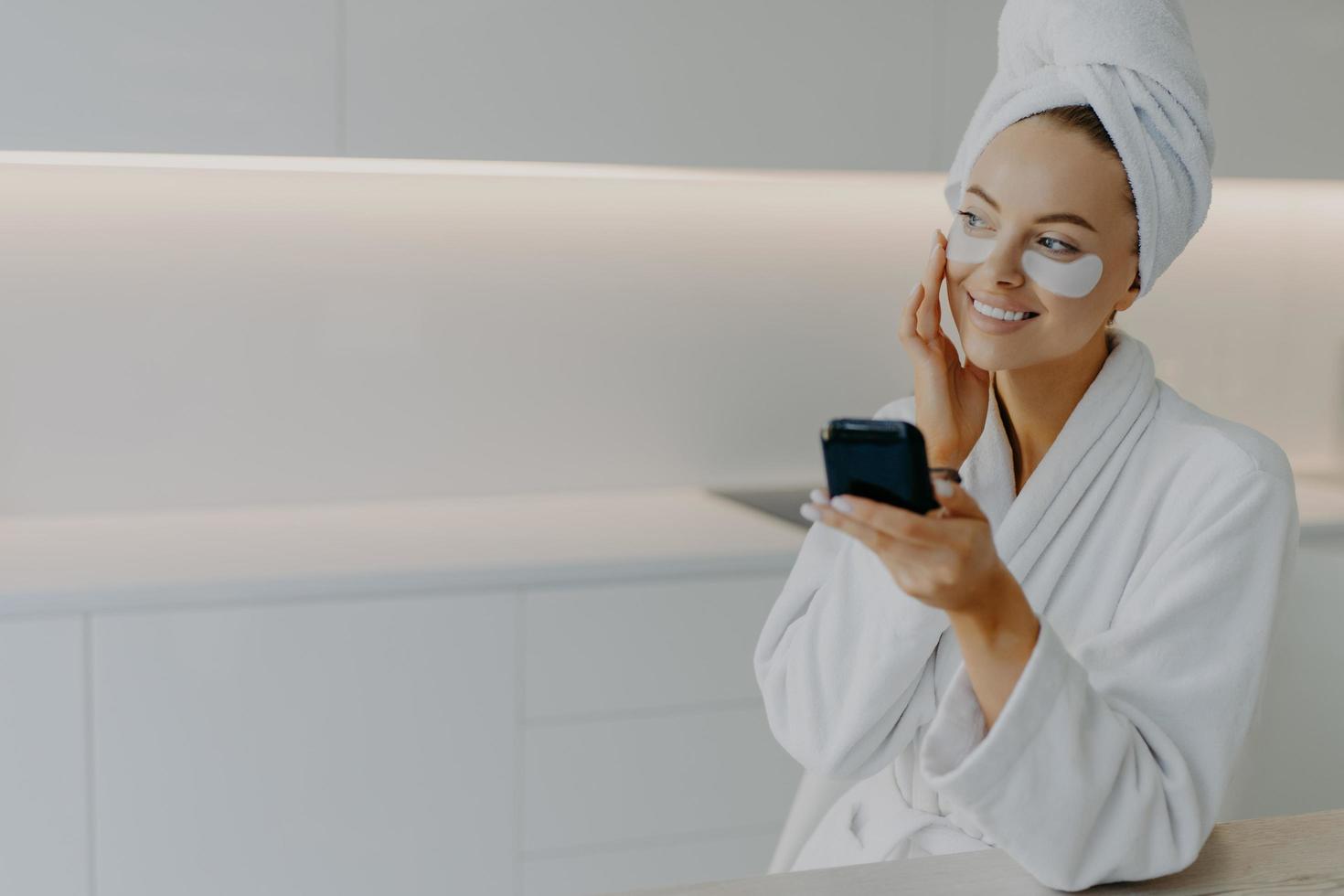 gelukkig glimlachen Europese vrouw is van toepassing hydraterend patches onder ogen accenten gezicht voorzichtig geniet huid zorg routine- gekleed in wit badjas looks ergens terzijde poses Bij keuken. schoonheid concept foto