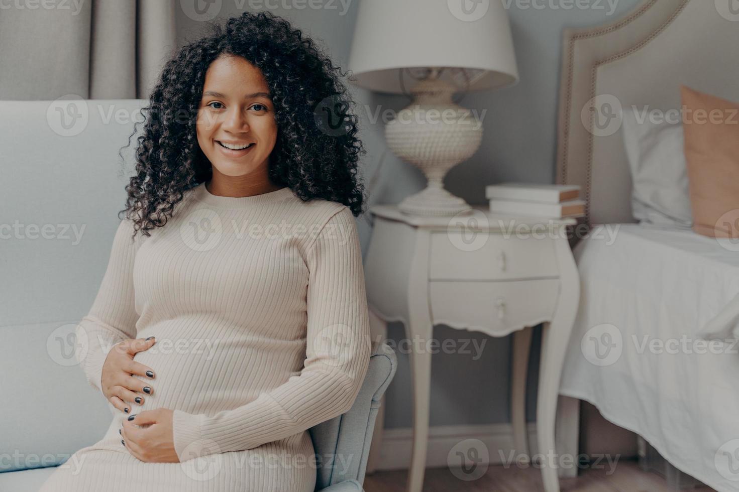 jonge gelukkige zwangere vrouw met krullend haar die haar buik vasthoudt en naar de camera glimlacht foto