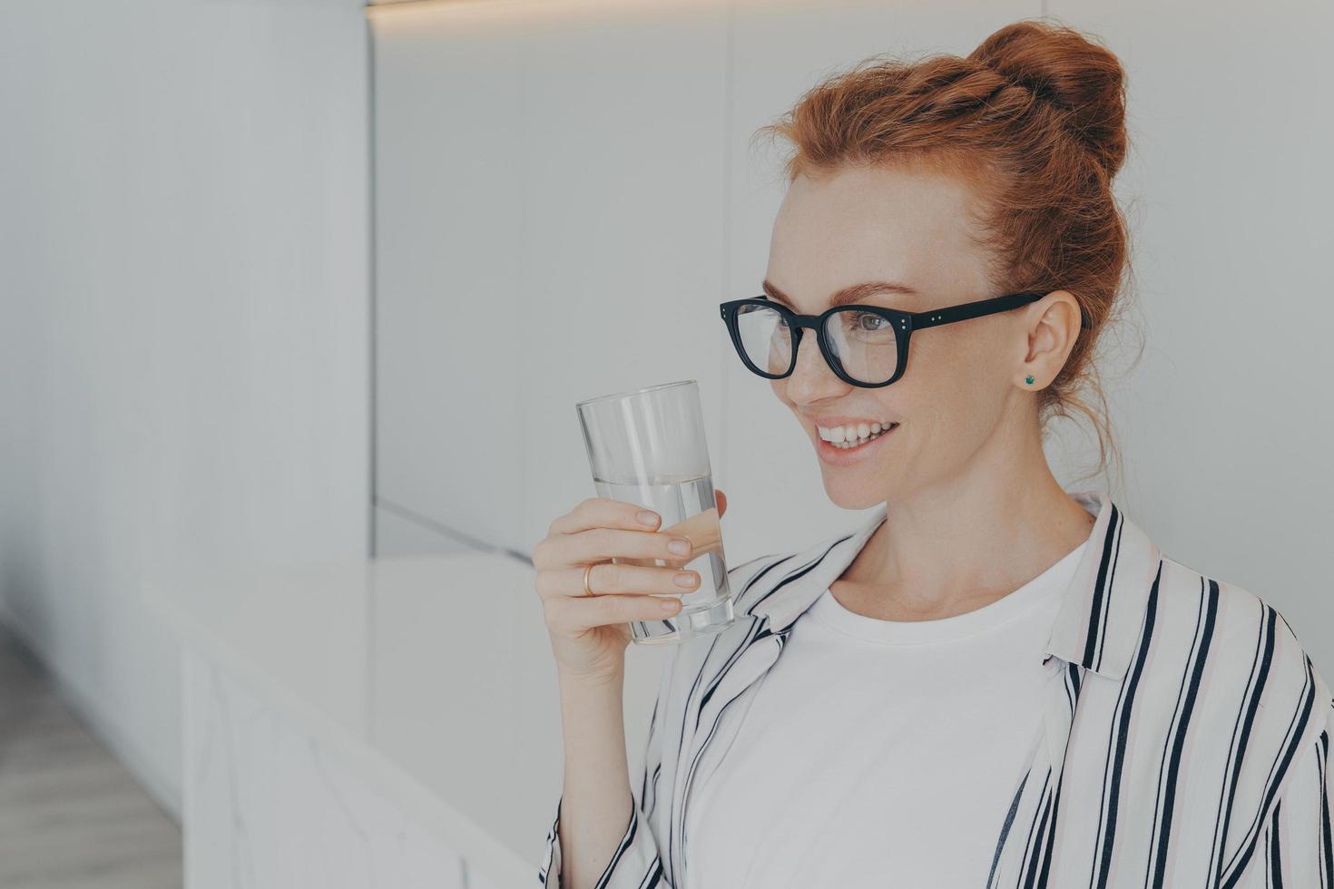 lachende roodharige jonge vrouw houdt glas water drinkt aqua voor lichaamsverfrissing foto
