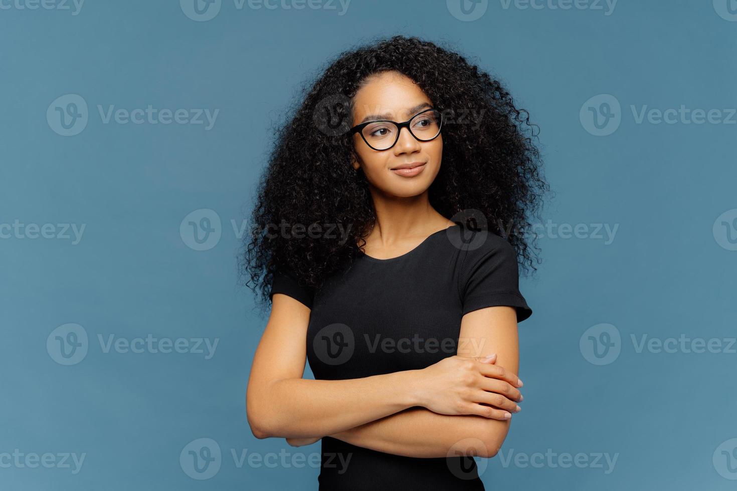 foto van doordachte tevreden afro-vrouw houdt handen gekruist over borst, gefocust opzij, draagt transparante bril, casual zwart t-shirt, geïsoleerd op blauwe achtergrond. menselijke gezichtsuitdrukkingen concept