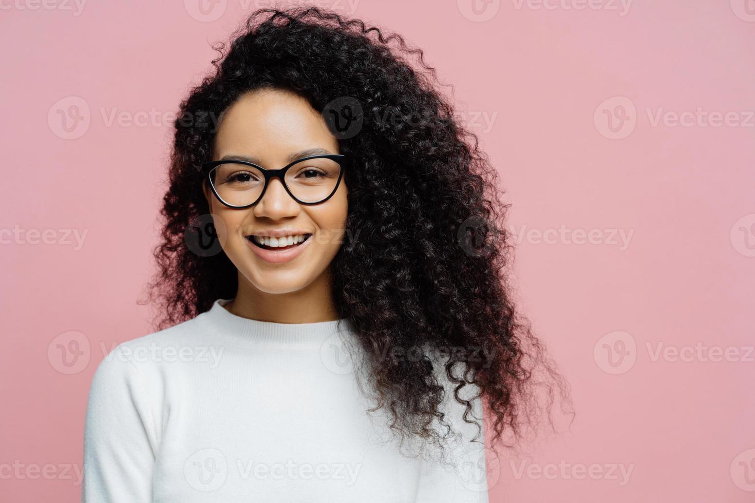 headshot van charmante jonge vrouw met afro kapsel, glimlacht aangenaam naar de camera, voelt zich erg gelukkig, draagt een transparante bril en witte trui, geïsoleerd op een roze achtergrond. geamuseerde etnische vrouw foto