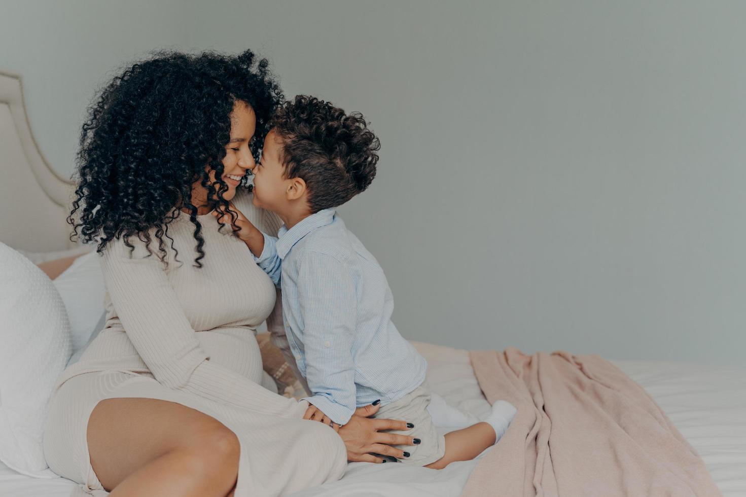 Afro-Amerikaanse moeder en zoontje zittend op bed en elkaar aanraken met neuzen foto