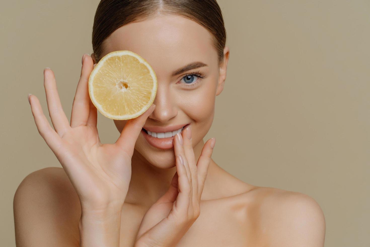 indoor shot van mooie vrouwelijke model heeft verse gezonde huid goed verzorgd lichaam dekt oog met schijfje sinaasappel poses blote schouders geïsoleerd over beige achtergrond. ingrediënt voor het maken van gezichtsmasker foto