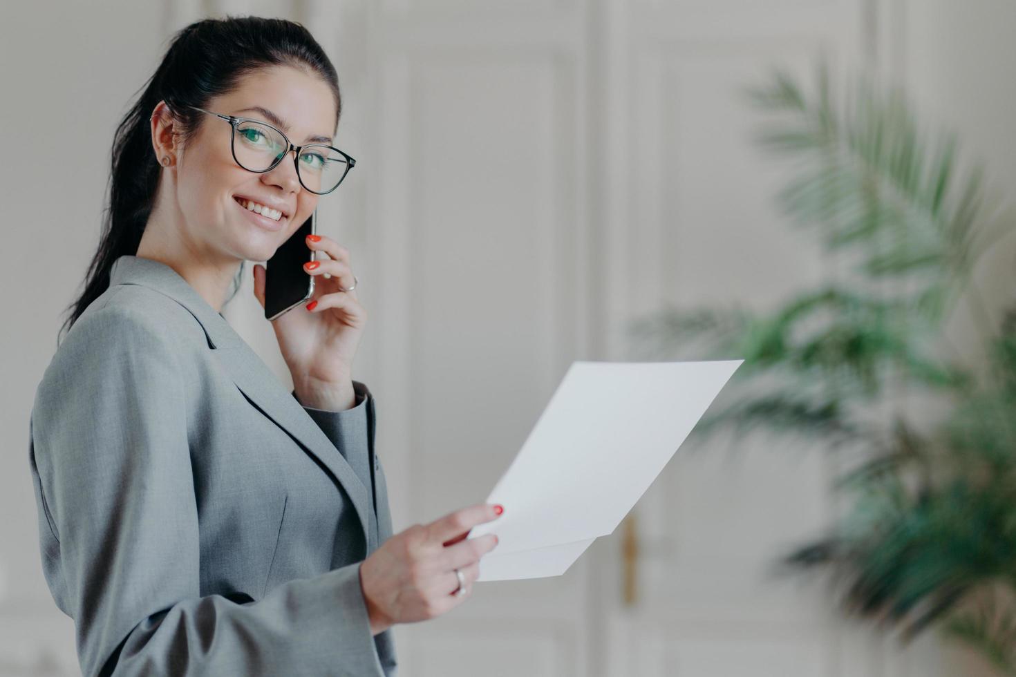 drukke vrouwelijke ondernemer heeft telefoongesprek, bezig met boekhoudkundig werk, kijkt door papieren documenten, controleert informatie, draagt bril en formele kostuum, poseert binnen, gezellig interieur foto