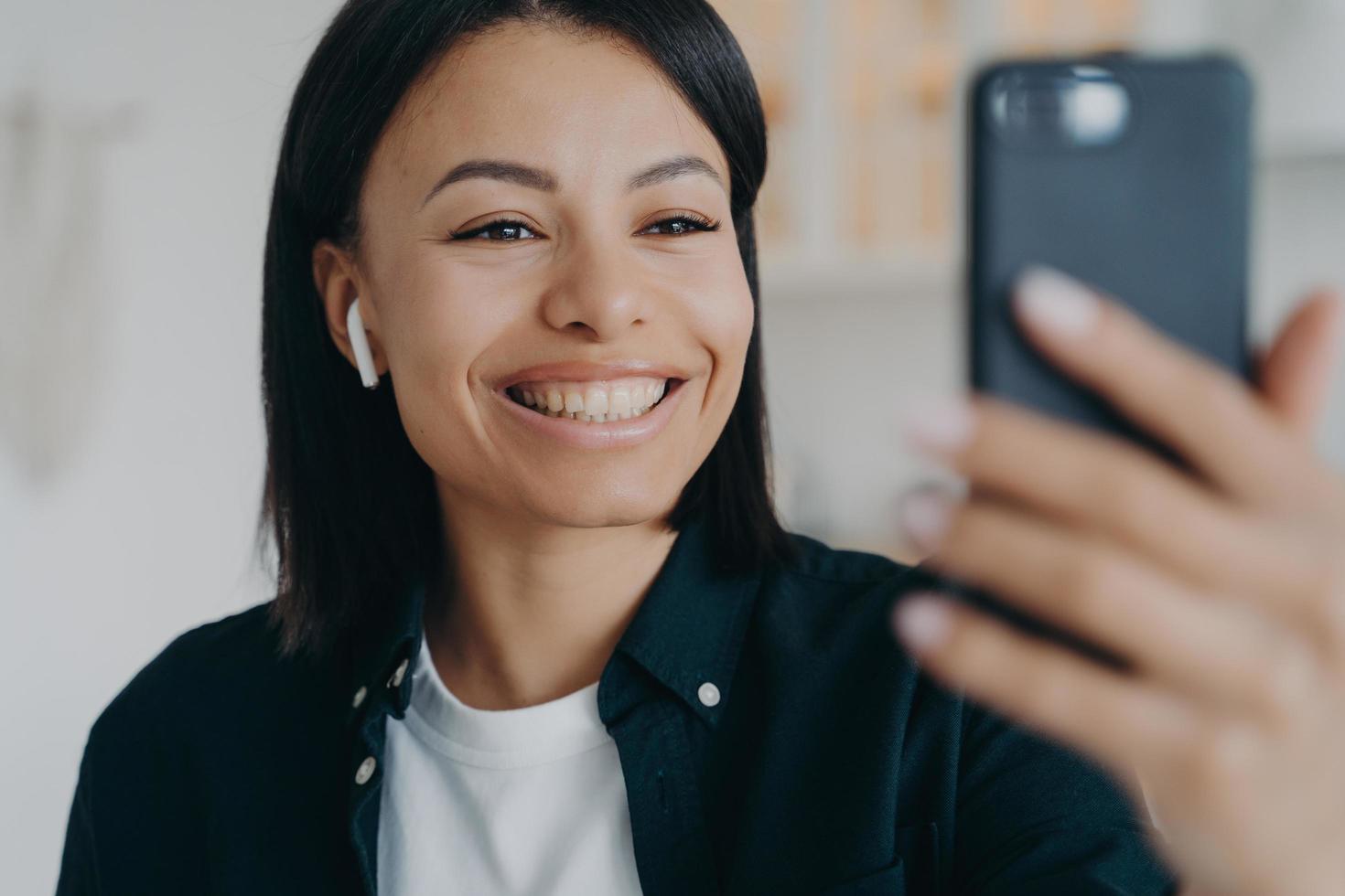 glimlachende vriendelijke jonge vrouw die een oortelefoon draagt, communiceert via een videogesprek met een smartphone in de hand foto