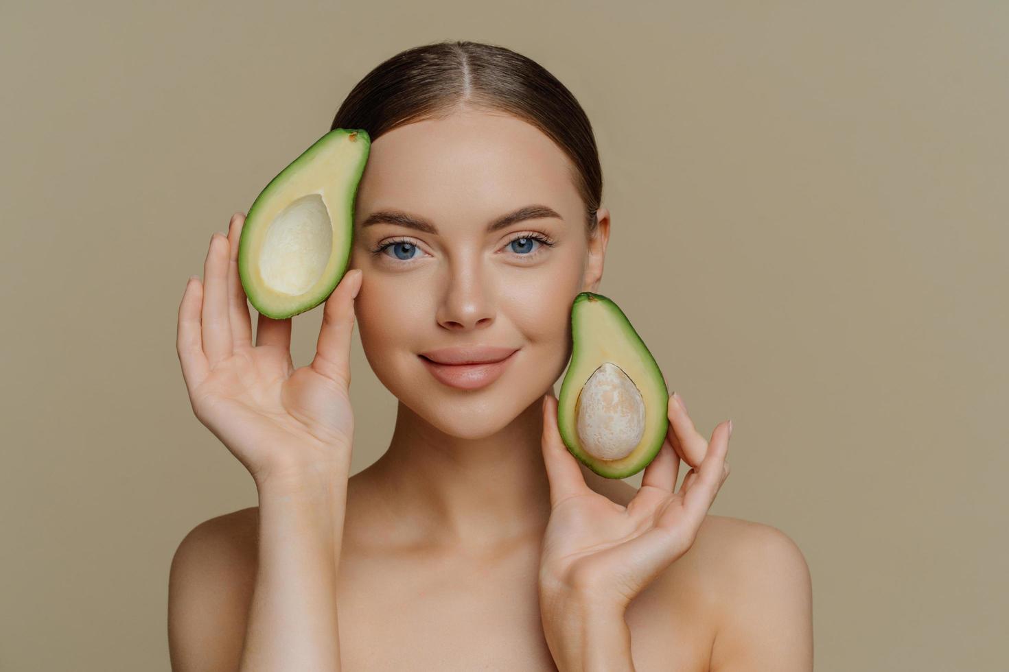 foto van zonder shirt vrouw model- looks met teder uitdrukking Bij camera houdt helften van avocado in de buurt gezicht heeft gezond goed verzorgd huid na kunstmatig behandelingen geïsoleerd over- beige achtergrond.