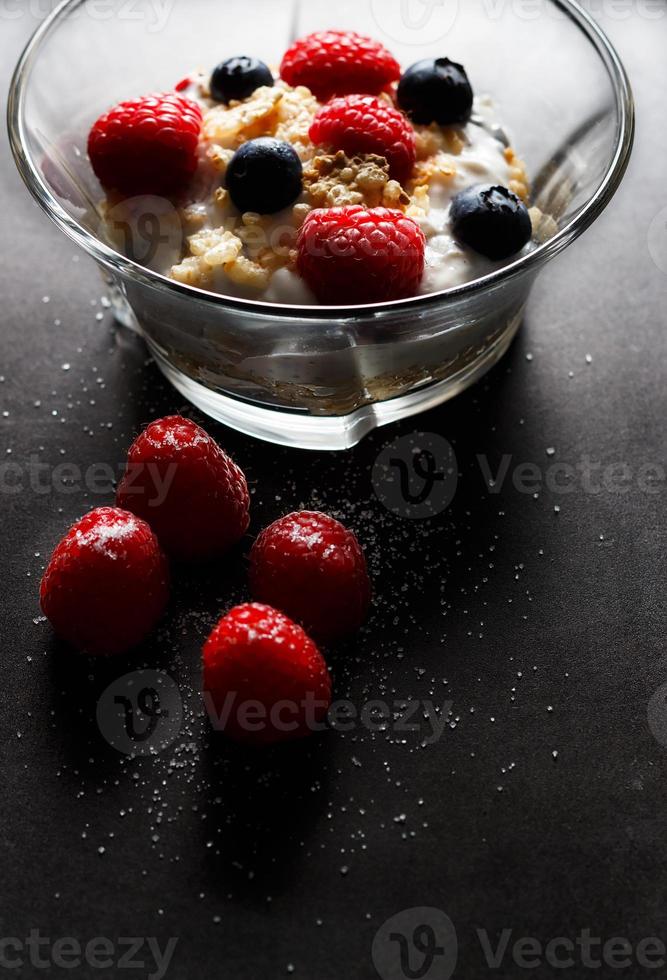 frambozen, bosbessen, granen en yoghurt in een glas kom Aan een zwart oppervlak. gezond ontbijt voor een gezond leven. verticaal afbeelding. foto