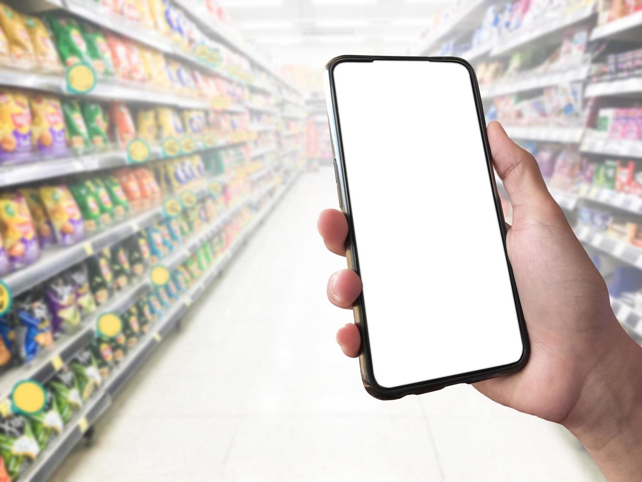 slim boodschappen doen concept mobiel telefoon in hand- in voorkant van goederen schappen in supermarkt en kruidenier op te slaan. blanco wit scherm mockup voor uw eigen creativiteit. foto