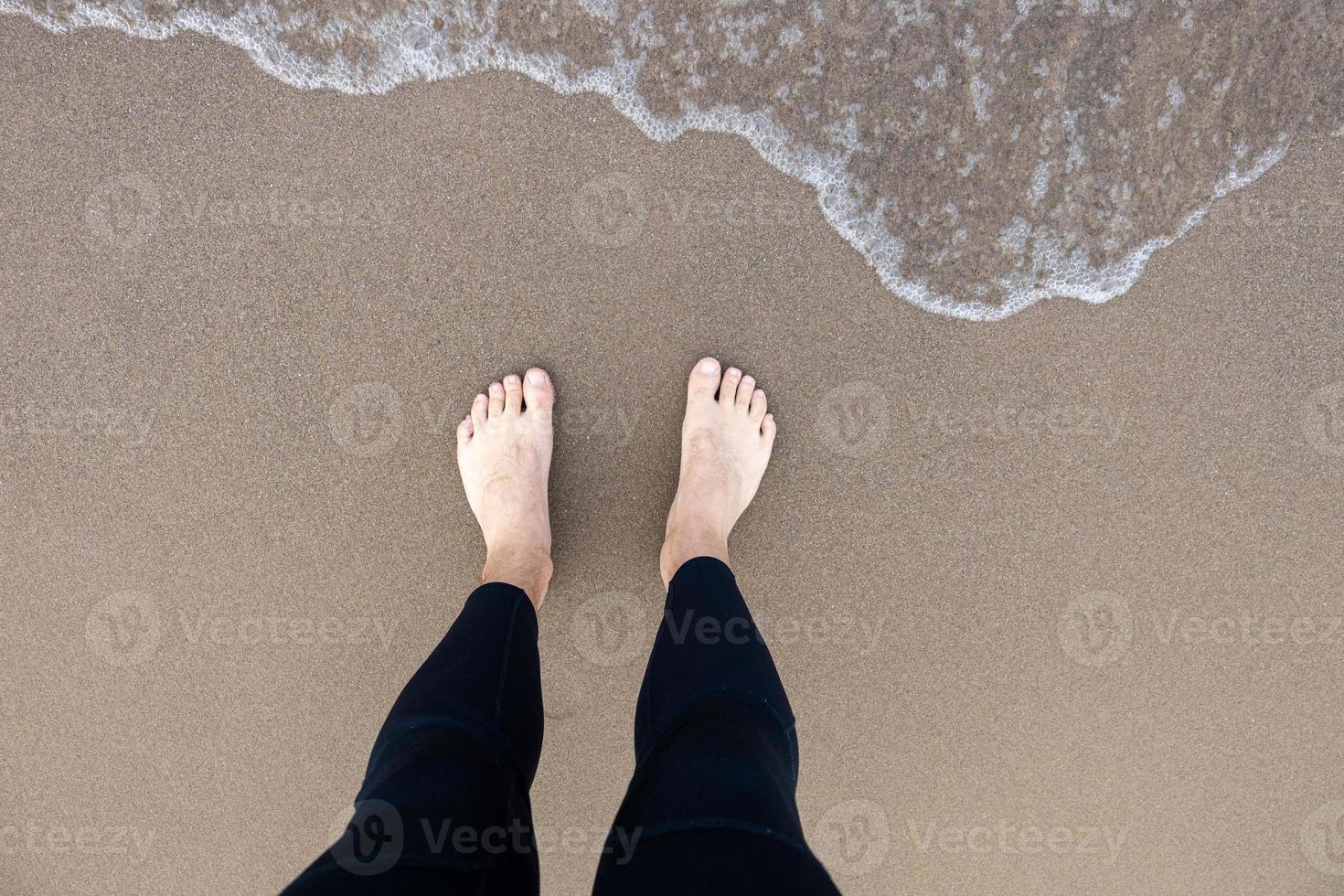 Mens vervelend een wetsuit staand Aan een schoon zanderig strand Aan meer Michigan aan het kijken de water komen naar zijn voeten foto