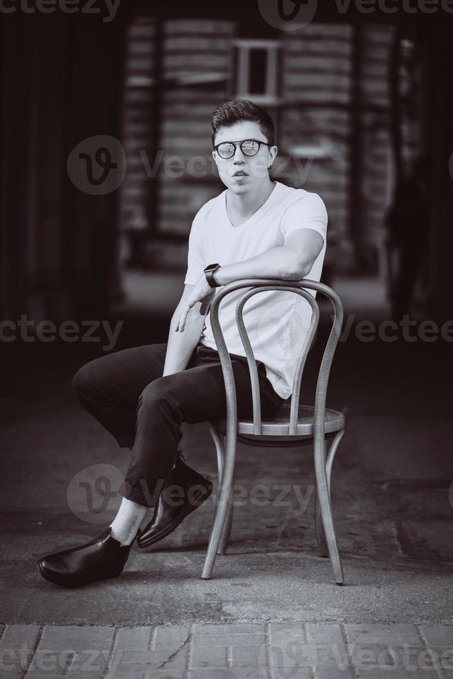 portret van mannen zittend Aan stoel met wit t-shirt en zonnebril in de straat foto