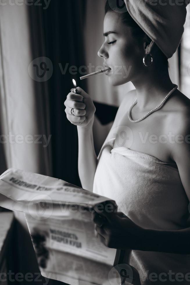 mooi jong vrouw in een handdoek rookt een sigaret en leest krant- foto