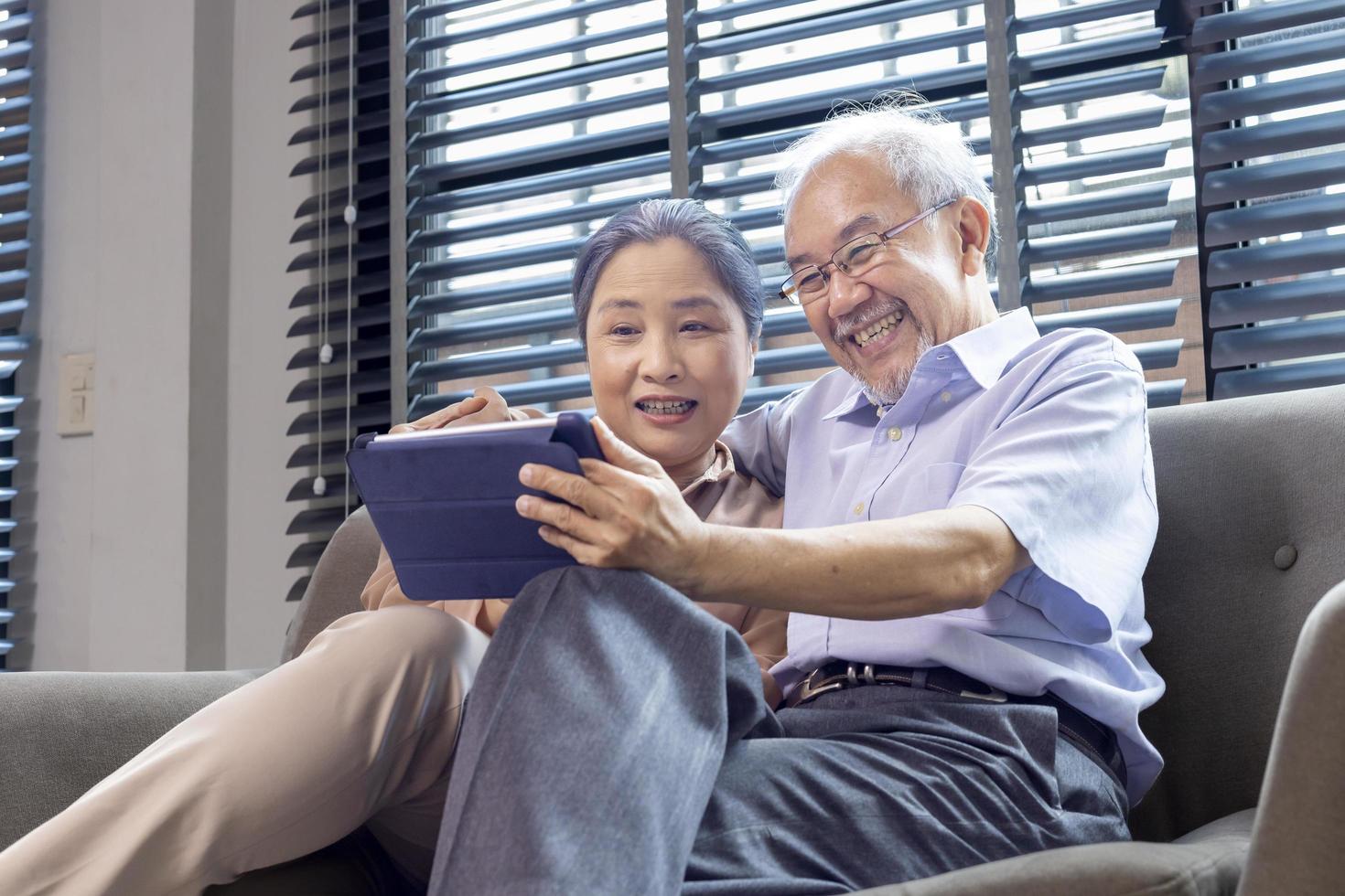 senior Aziatisch paar in pensioen leeftijd op zoek Bij nostalgisch foto gebruik makend van digitaal tablet terwijl zittend Aan de sofa bankstel in hun pensioen huis voor ouderling en pensioen activiteiten concept
