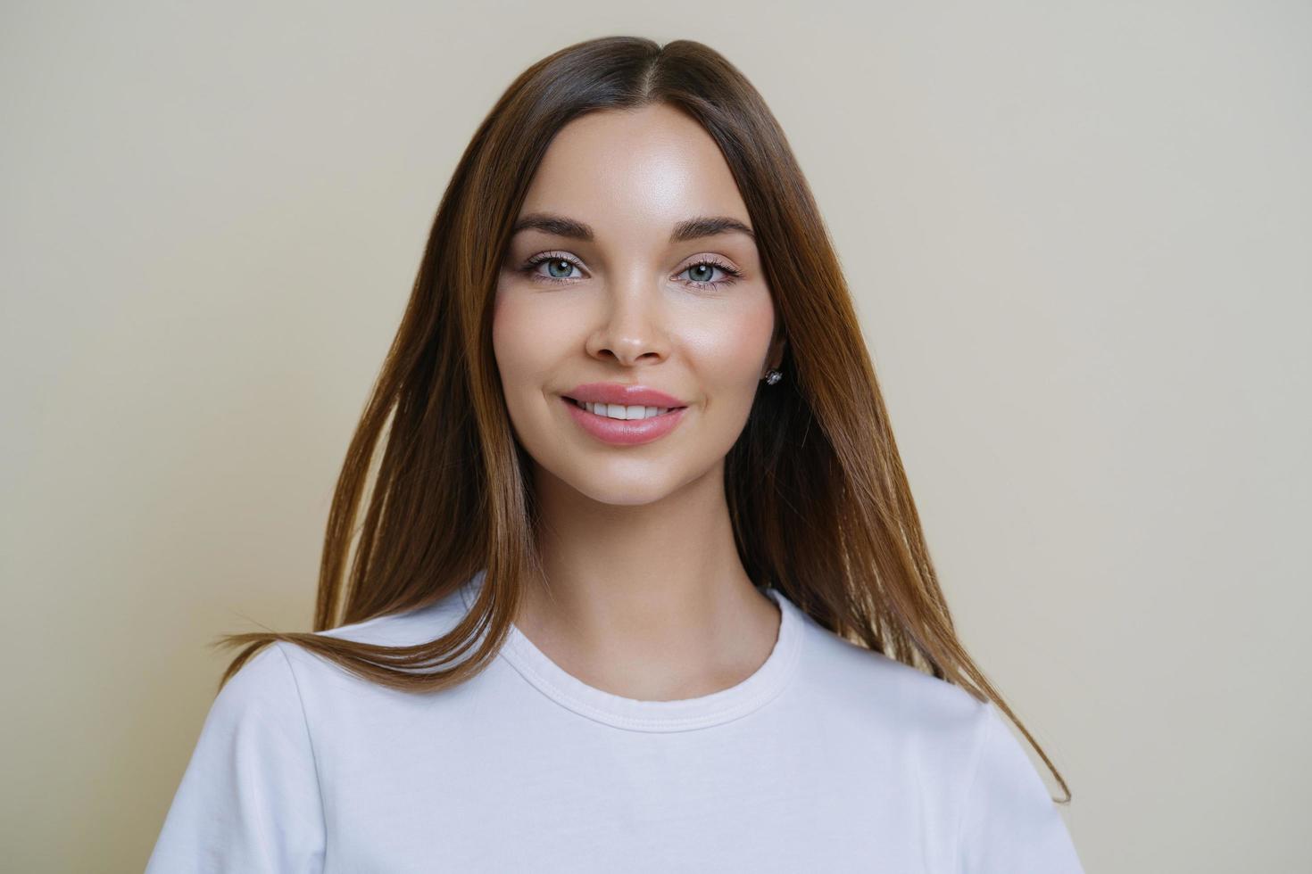 headshot van mooie donkerharige jonge Europese vrouw kijkt met tedere uitdrukking, heeft een vriendelijke houding, praat nonchalant met vrienden, draagt een casual wit t-shirt, geïsoleerd op beige achtergrond foto