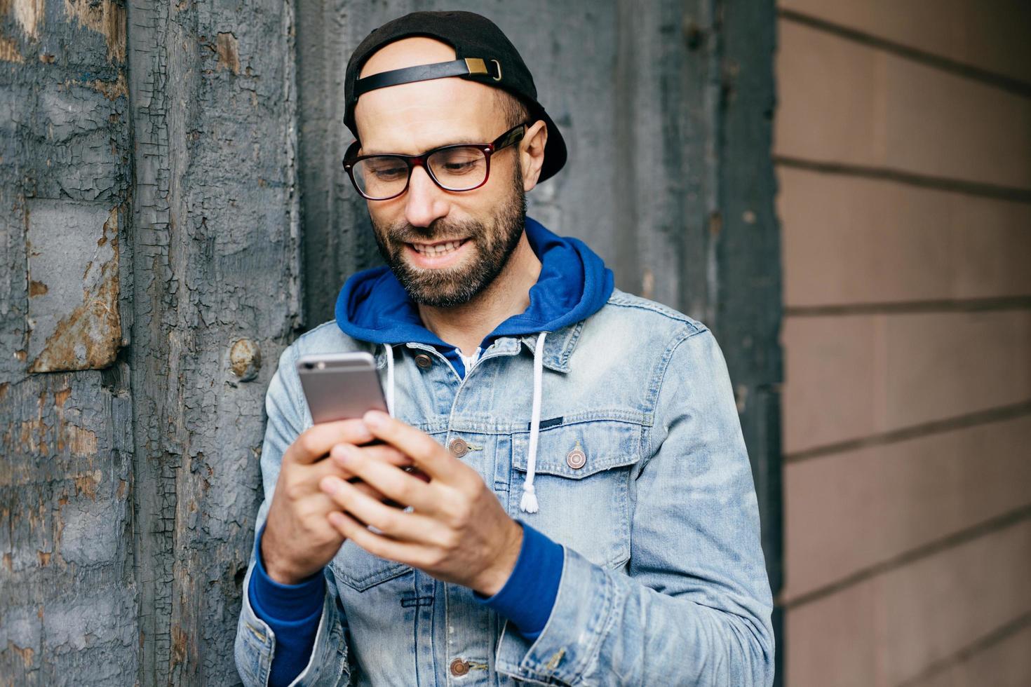 horizontaal portret van een knappe, stijlvolle man met een baard in een denim anorak met een grote bril die een smartphone vasthoudt met behulp van gratis internet, e-mail checkt, grappige video's kijkt, applicaties updatet foto