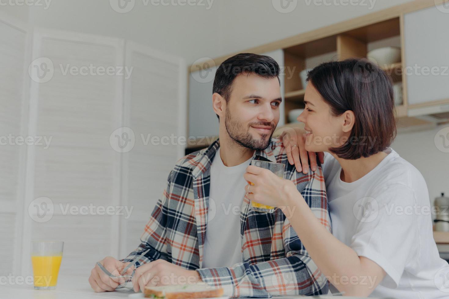 mooi stel kijkt elkaar liefdevol aan, vrouw houdt een glas sap vast, poseert samen bij de keukentafel, praat gezellig tijdens het ontbijt, geniet van de huiselijke sfeer. gelukkig familieconcept foto