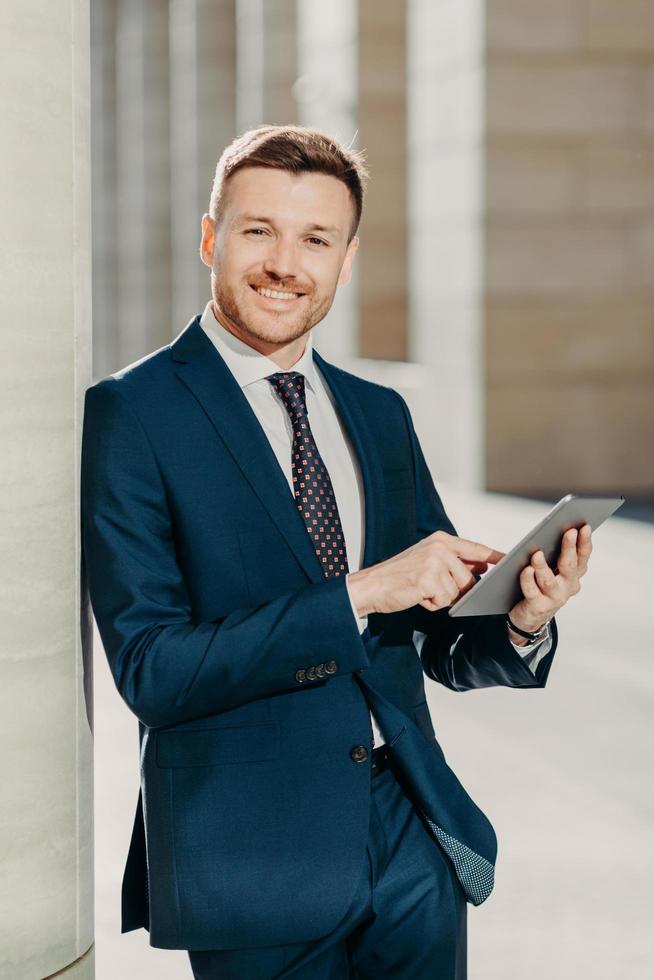 verticaal schot van zakenman gebruikt digitale tablet voor online communicatie en werk, maakt gebruik van gratis internet op kantoor, heeft positieve expressie, gekleed in formeel zwart pak. mensen en beroep foto