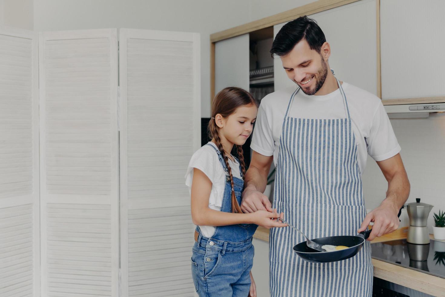 klein vrouwelijk kind helpt haar vader om het ontbijt te bereiden, samen eieren te bakken, te genieten van de huiselijke sfeer, in de keuken te staan, vader draagt een schort en houdt een koekenpan vast. familie en kooktijd concept foto