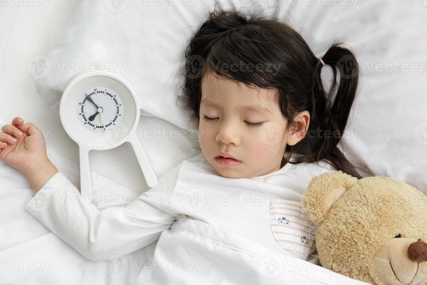 kind meisje slapen met teddy beer Aan de houten bed in haar slaapkamer, gelukkig Aziatisch kind weinig meisje en alarm klok wakker worden omhoog in de ochtend- foto