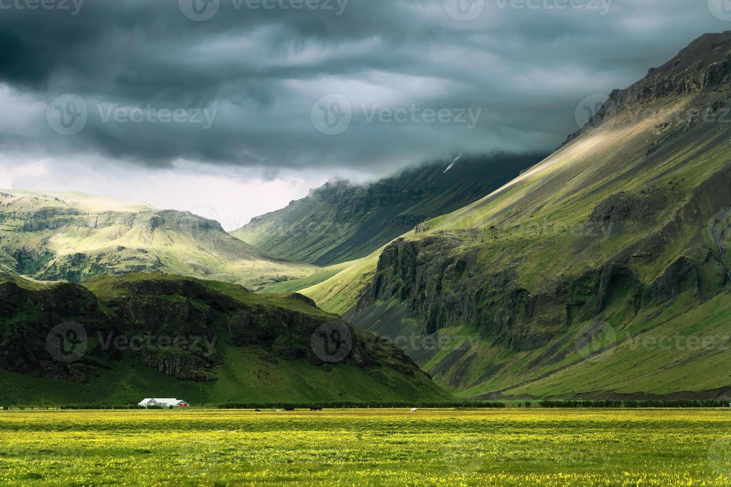 dramatisch IJslands berg met zonlicht schijnend door storm wolken en huis Aan veld- in zomer Bij IJsland foto