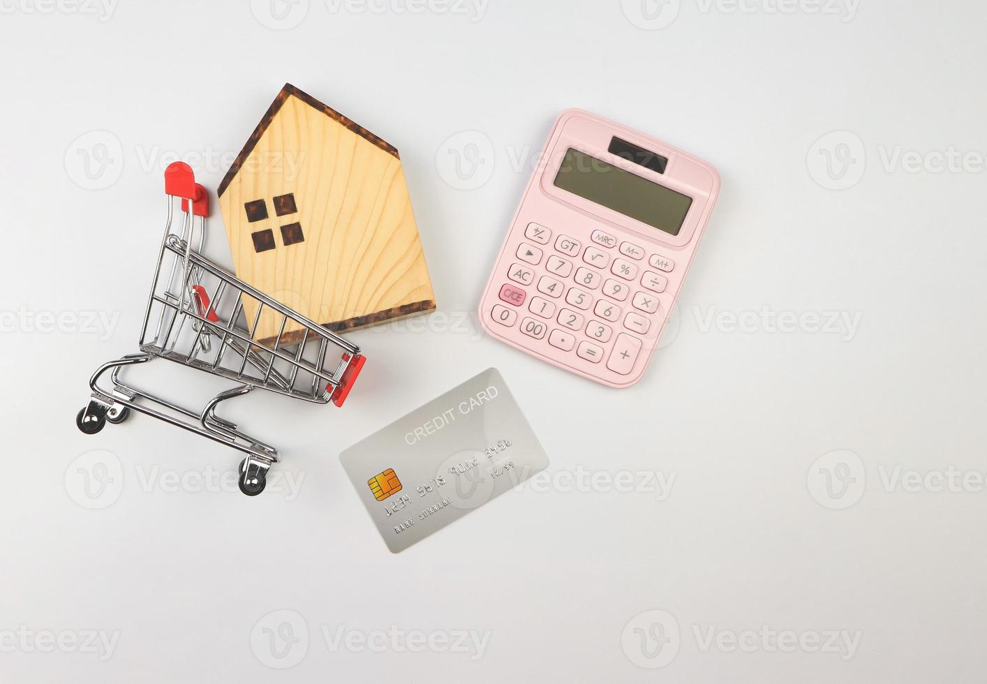 vlak lay-out van houten huis model- in boodschappen doen karretje, roze rekenmachine en credit kaart Aan wit achtergrond, huis aankoop berekening concept. foto