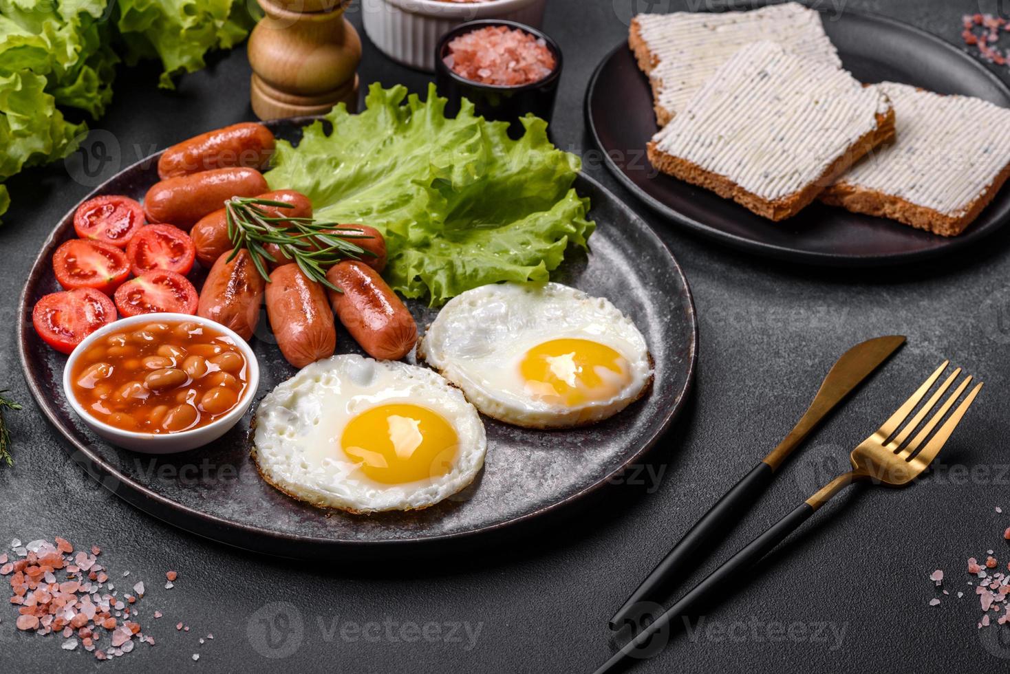 traditioneel Engels ontbijt met eieren, geroosterd brood, worstjes, bonen, specerijen en kruiden Aan een grijs keramisch bord foto