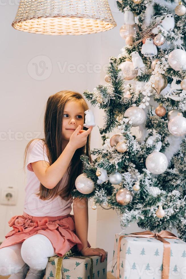 aanbiddelijk weinig meisje decoreren een Kerstmis boom met kerstballen Bij huis foto