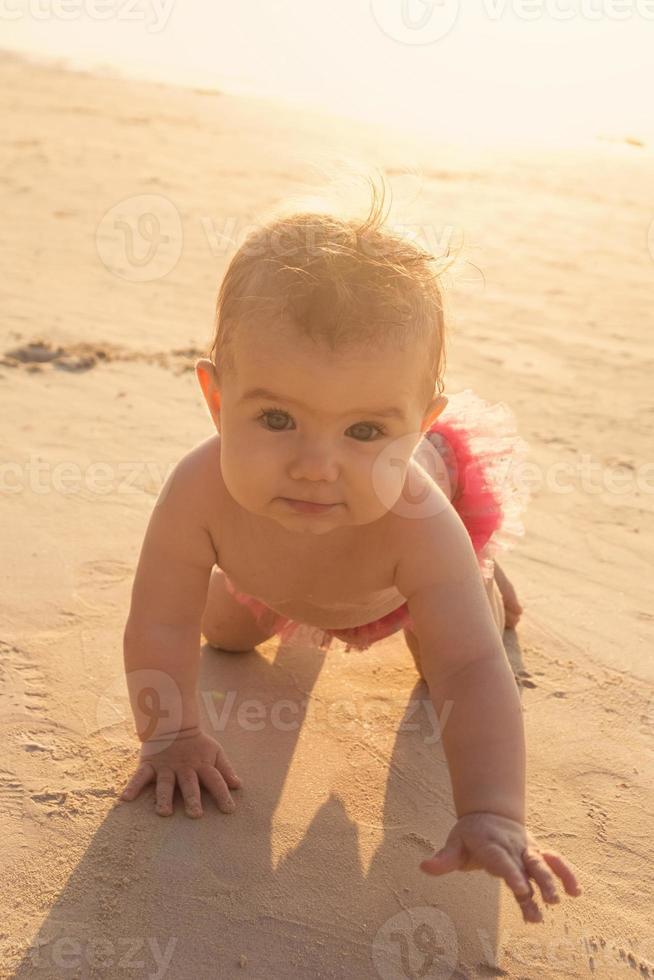 weinig baby meisje is kruipen Aan een zanderig strand in de buurt naar zee in zonsondergang zonlicht. foto