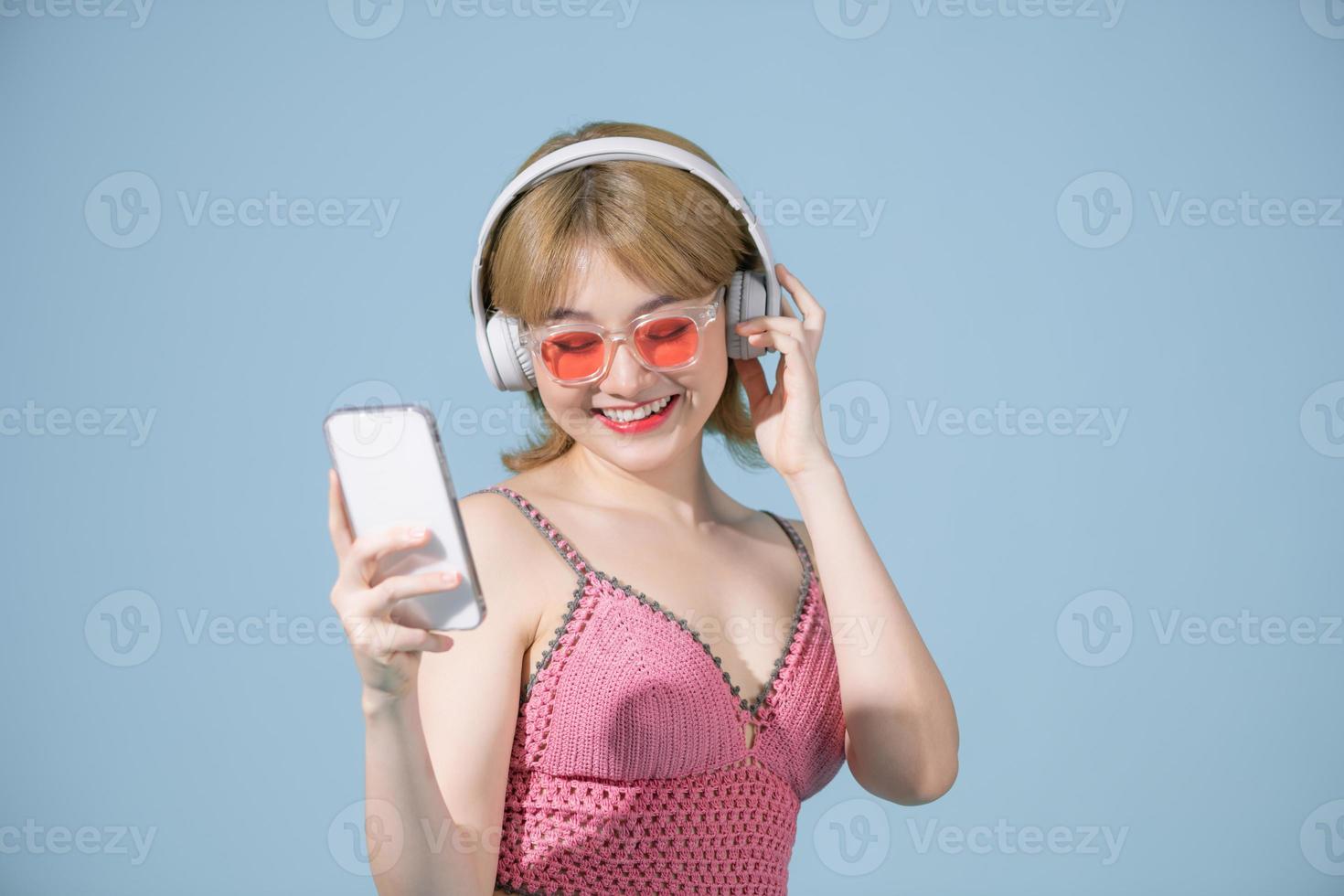 foto van opgewonden vrouw in badmode geïsoleerd over- blauw muur achtergrond tonen Scherm van mobiel telefoon luisteren muziek- met koptelefoon.