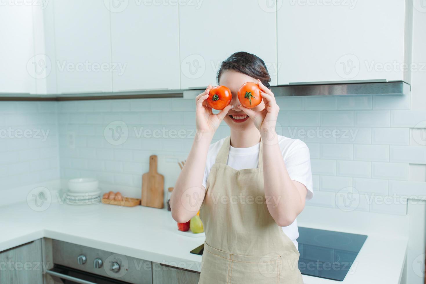 gelukkig humeur Aziatisch Dames Holding rood tomaten in de buurt de ogen, staand in de keuken, foto