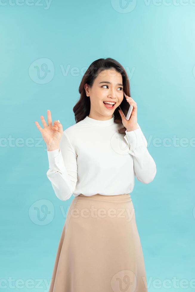 foto van charmant jong vrouw maken Oke gebaar terwijl pratend door haar telefoon over- blauw achtergrond. kijken Bij camera.
