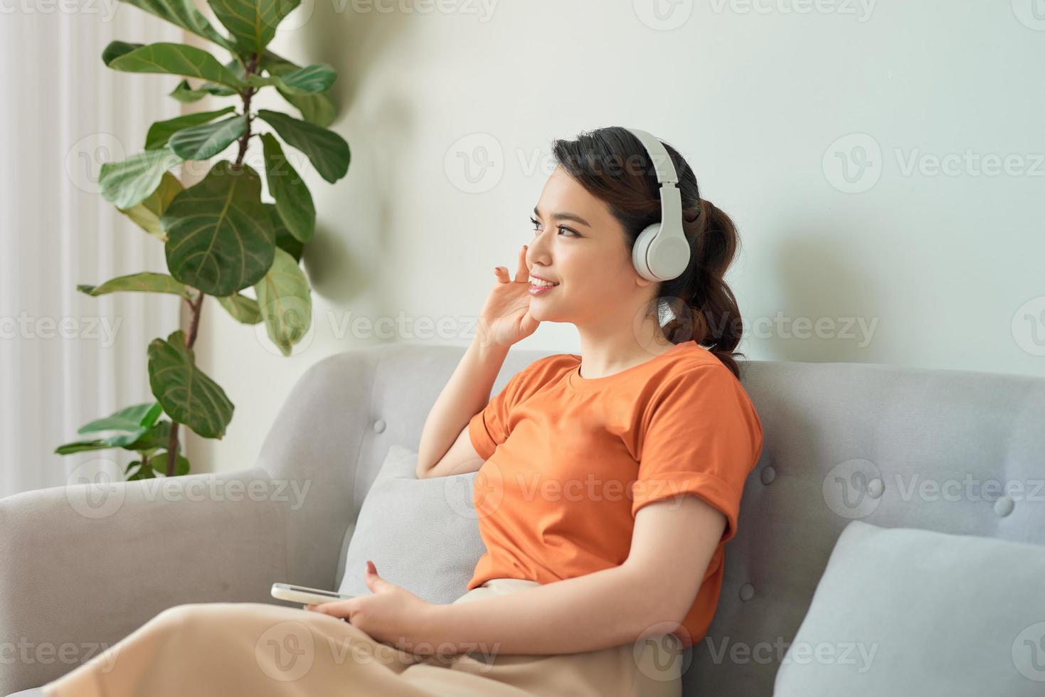 verheugd jong vrouw luisteren naar muziek- met hoofdtelefoons en gebruik makend van mobiel telefoon terwijl leunend Aan een bankstel Bij huis foto