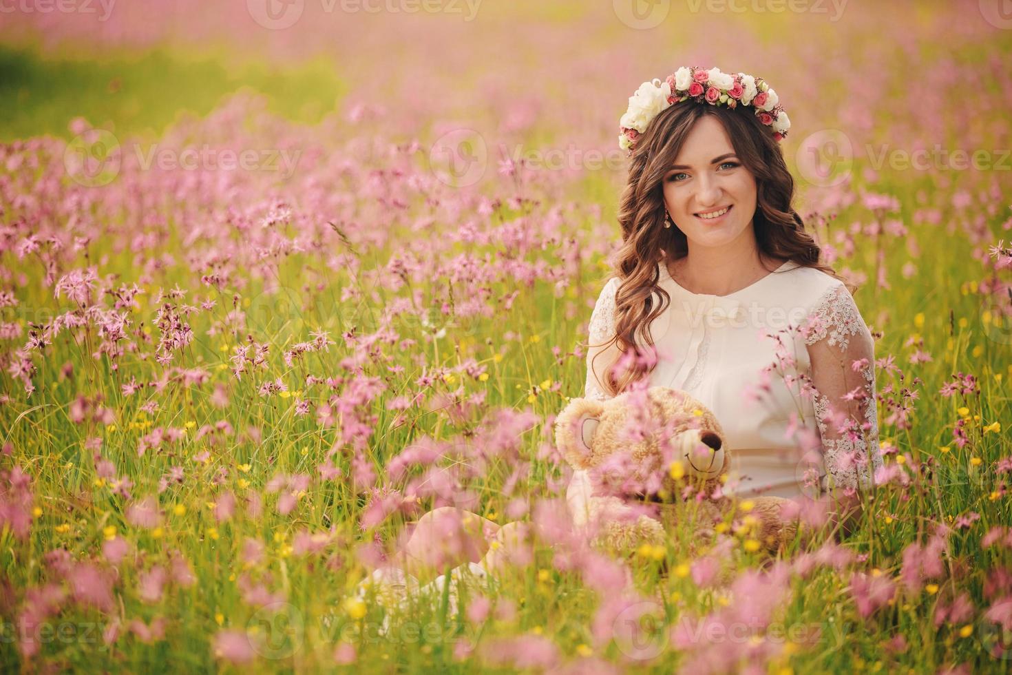 portret van een zwanger vrouw met teddy beer in fieid van roze bloemen. jong mooi zwanger meisje met een krans Aan haar hoofd in de zon. moederschap. de lente. kopiëren ruimte. selectief focus foto