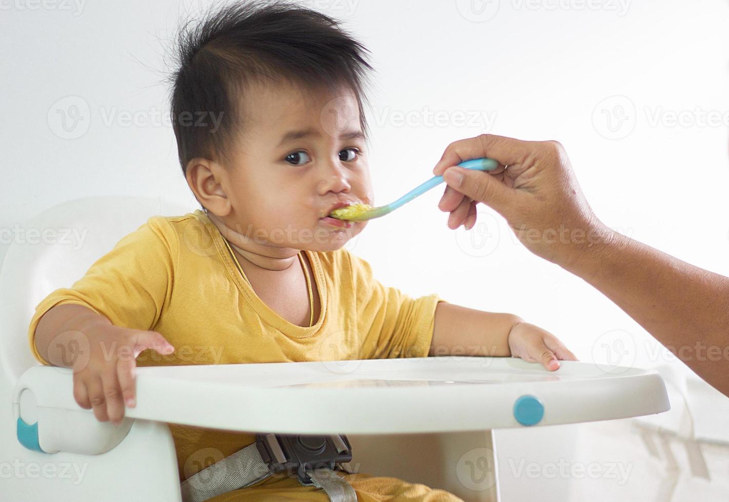 schattig wit meisje zittend stoel aan het eten ontbijt in binnen- huis voeden heerlijk maaltijden dat zijn zeer voedzaam de baby heeft een vrolijk gezicht grappig glimlachen en gelukkig in een gezond familie levensstijl. foto