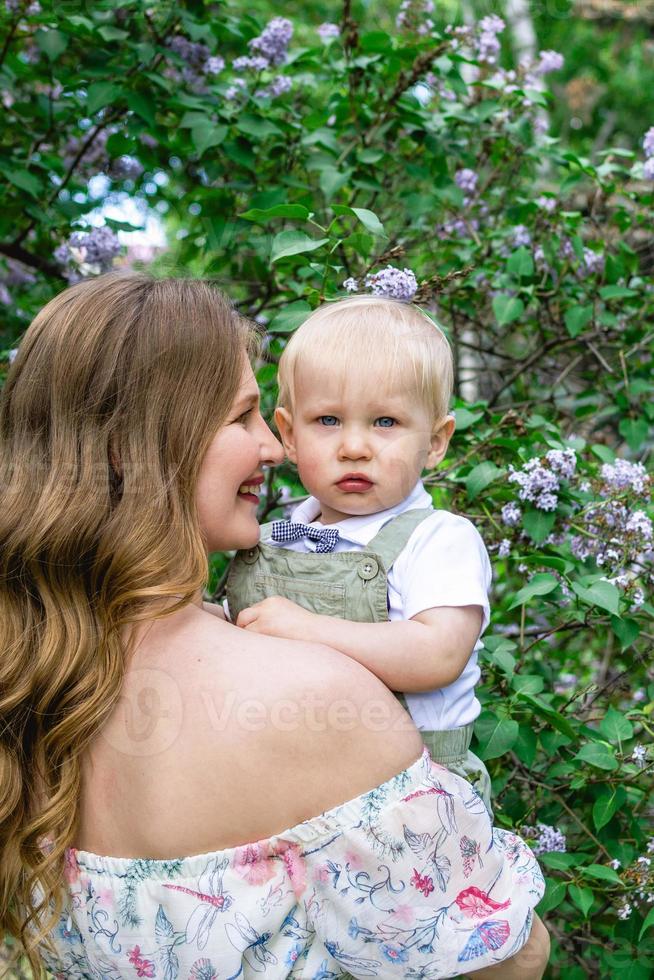 openhartig portret van jong glimlachen moeder met haar weinig zoon in tuin met bloeiend lila bloemen. foto