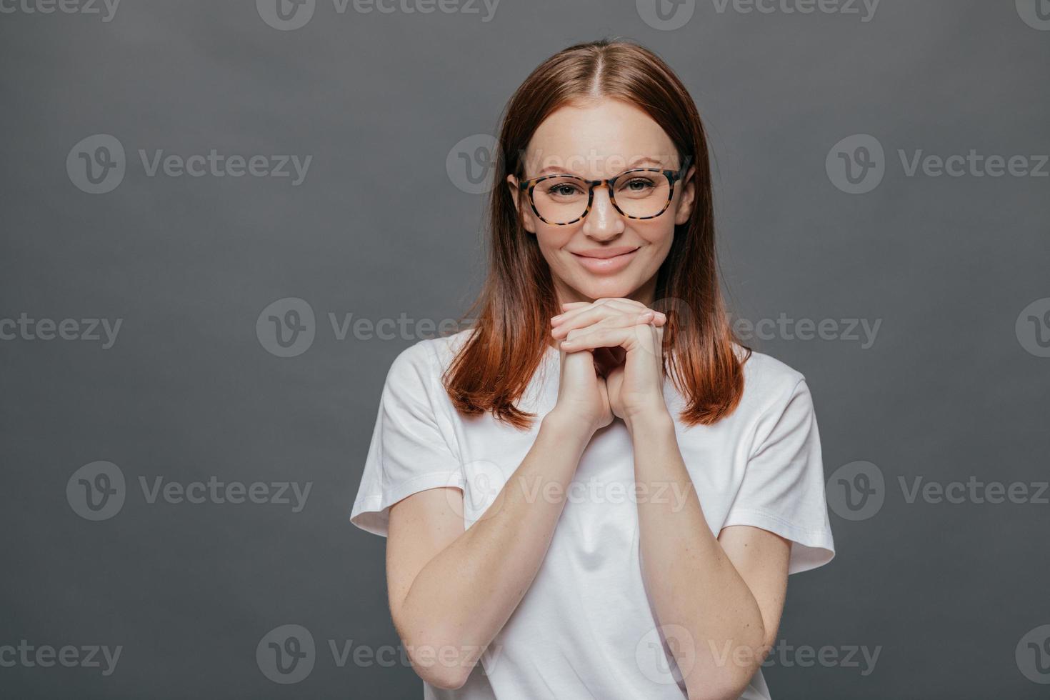 vrolijke aangenaam ogende dame houdt beide handen onder de kin, glimlacht positief naar de camera, draagt een optische bril, casual t-shirt, geniet van vrije tijd in goed gezelschap, geïsoleerd over grijze achtergrond foto