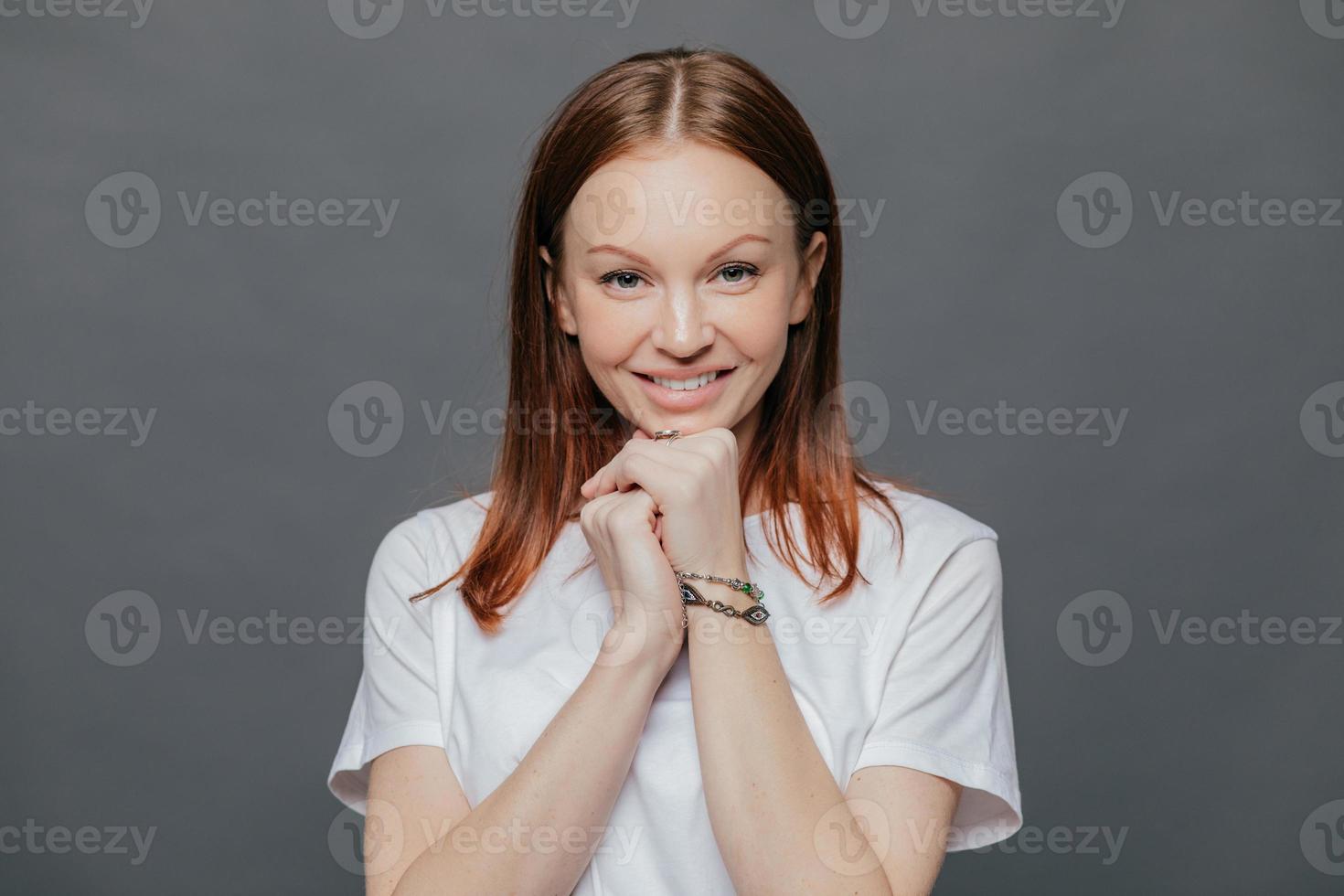 horizontaal schot van tevreden mooie vrouw draagt casual wit t-shirt, houdt handen onder de kin, glimlacht zachtjes, is in een goed humeur, poseert tegen donkergrijze studio-achtergrond. positieve emoties foto