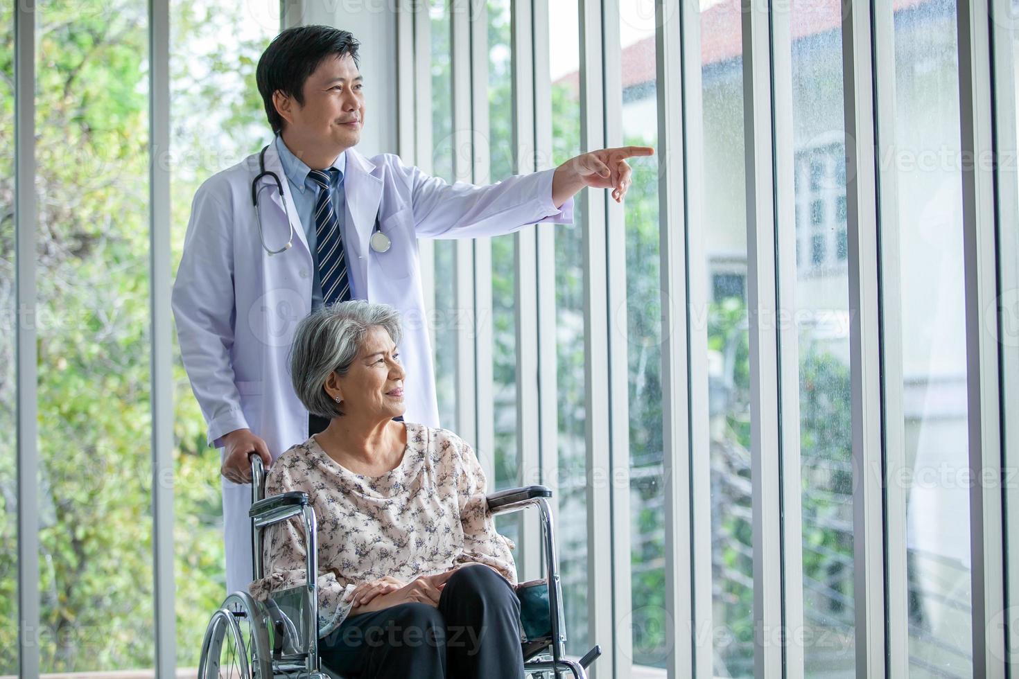 Aziatisch dokter ondersteunen en juichen omhoog senior geduldig in rolstoel praten, glimlachen in comfort Bij huis. gezondheidszorg en geneeskunde concept. foto