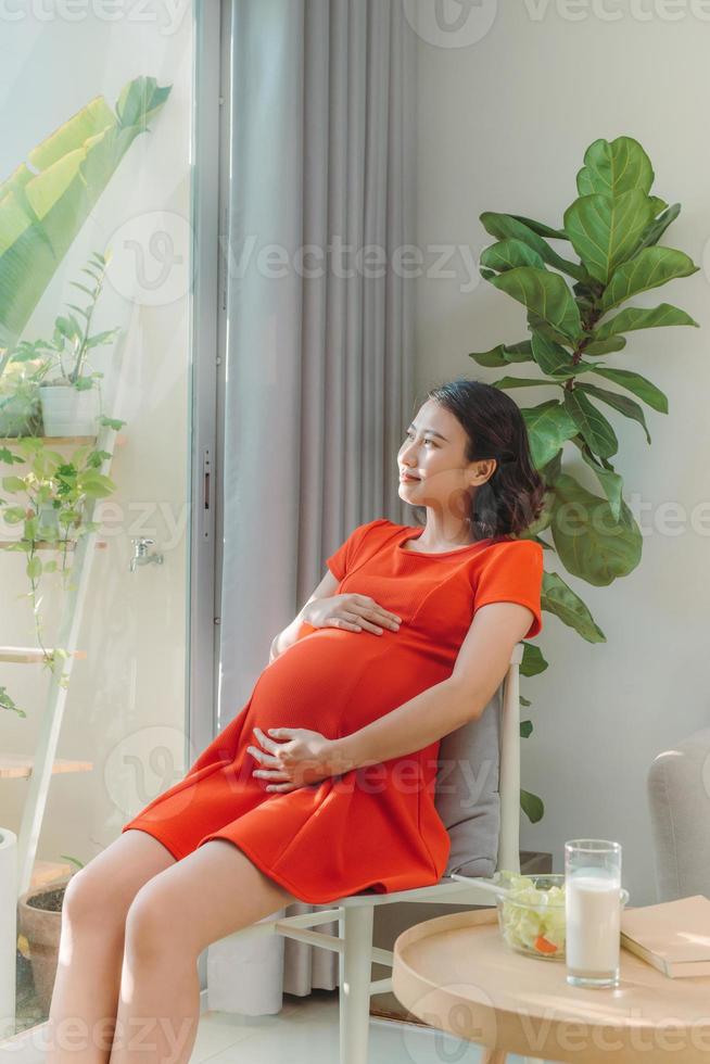 trots zwanger vrouw op zoek haar buik zittend Aan een sofa in de leven kamer Bij huis met een warm licht komt eraan in door de venster foto