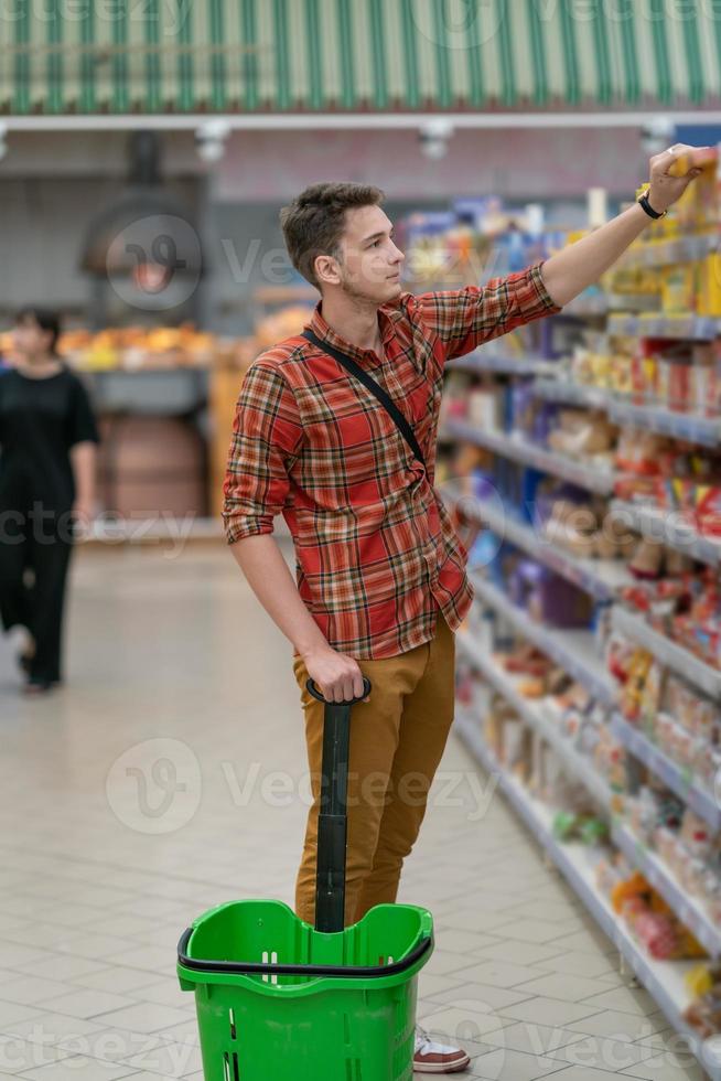 een jong Mens in een plaid overhemd koopt boodschappen in een supermarkt. een Mens duurt goederen van een plank in een op te slaan foto