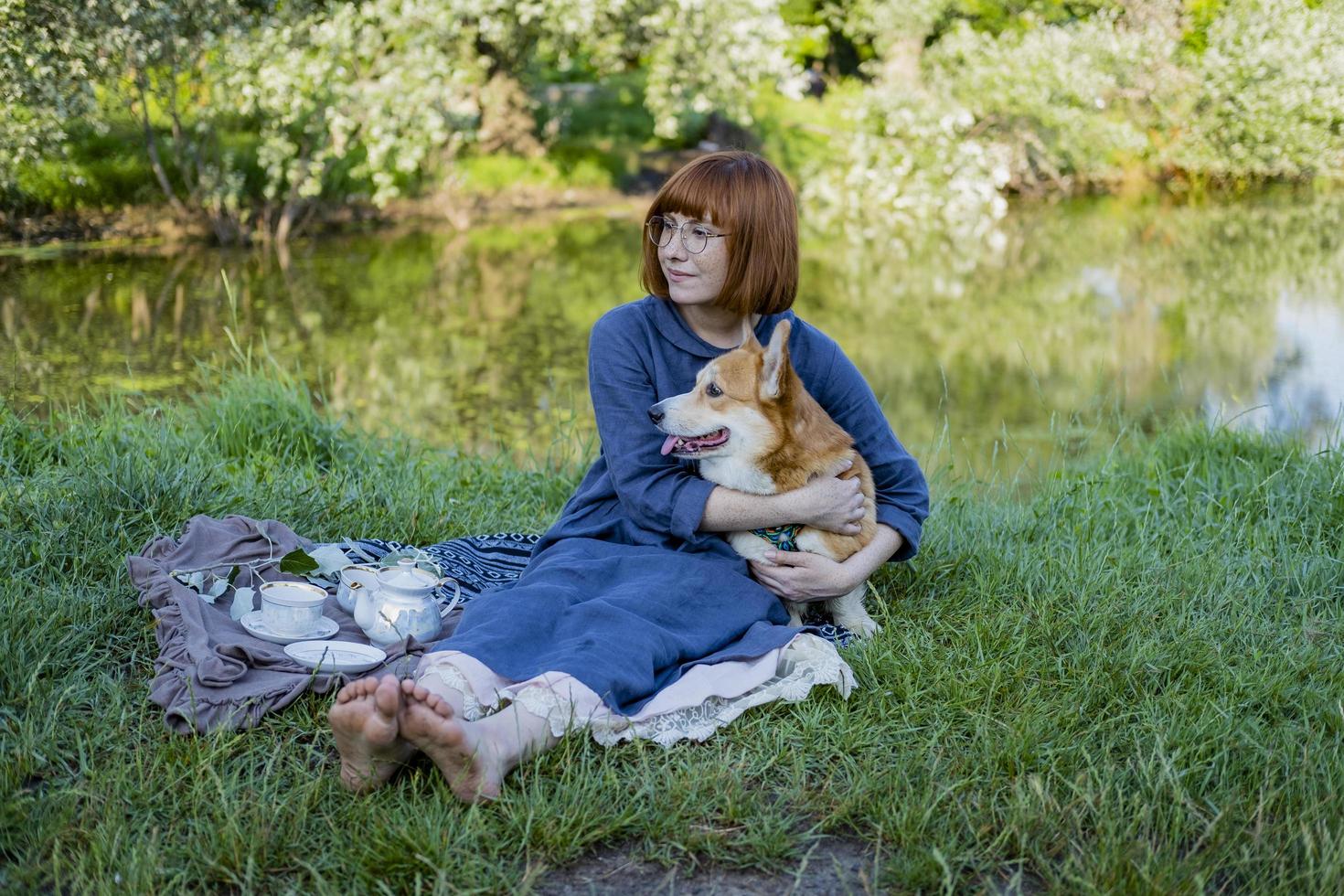jonge vrouw in retro jurk met grappige corgi-hond op de picknick, vrouw met schattige hond drinkt Engelse thee in het park foto