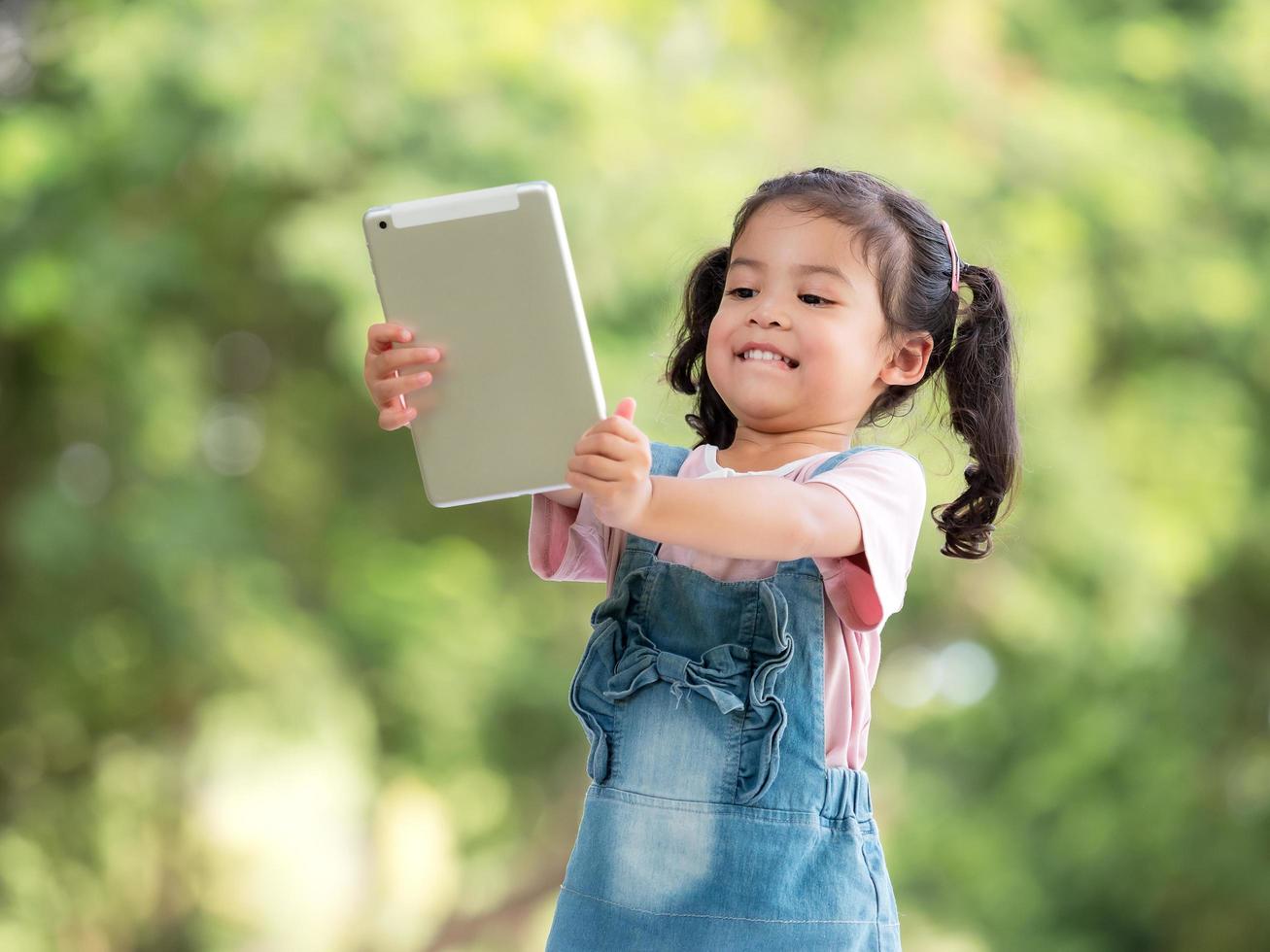 een schattig Aziatisch meisje gebruikt een tablet om spelletjes te spelen en buiten school in het park te leren foto