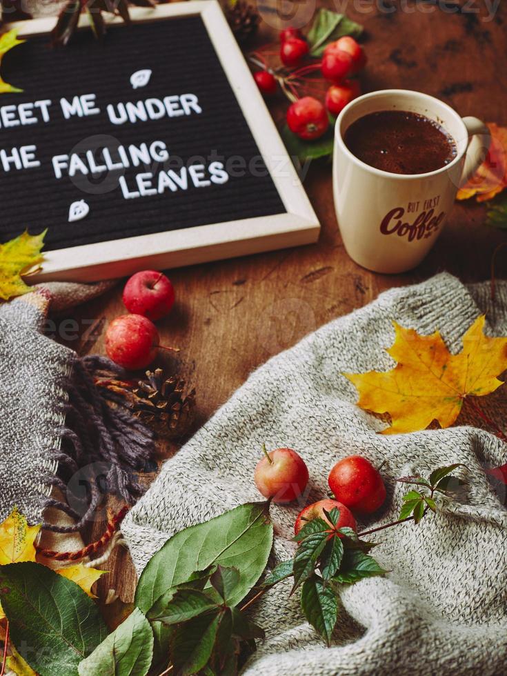 herfst flatlay met brief bord, knus trui en sjaal, kop van koffie, kleurrijk bladeren en klein appels Aan houten achtergrond foto